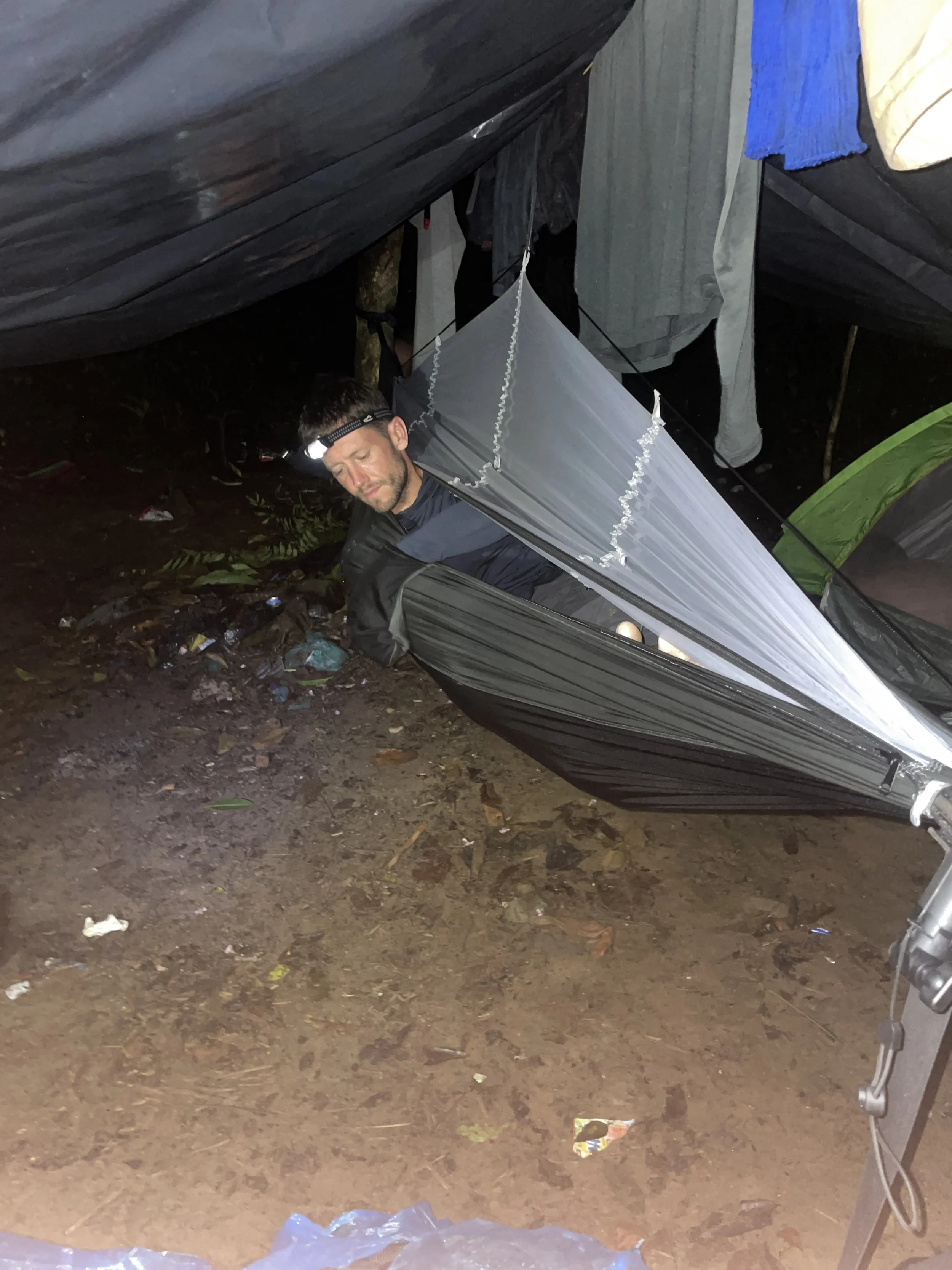 A man lying in a black and gray hammock suspended between trees in a wooded area at night, wearing a headlamp and sleeping gear. Clothes are hanging from a line above him.