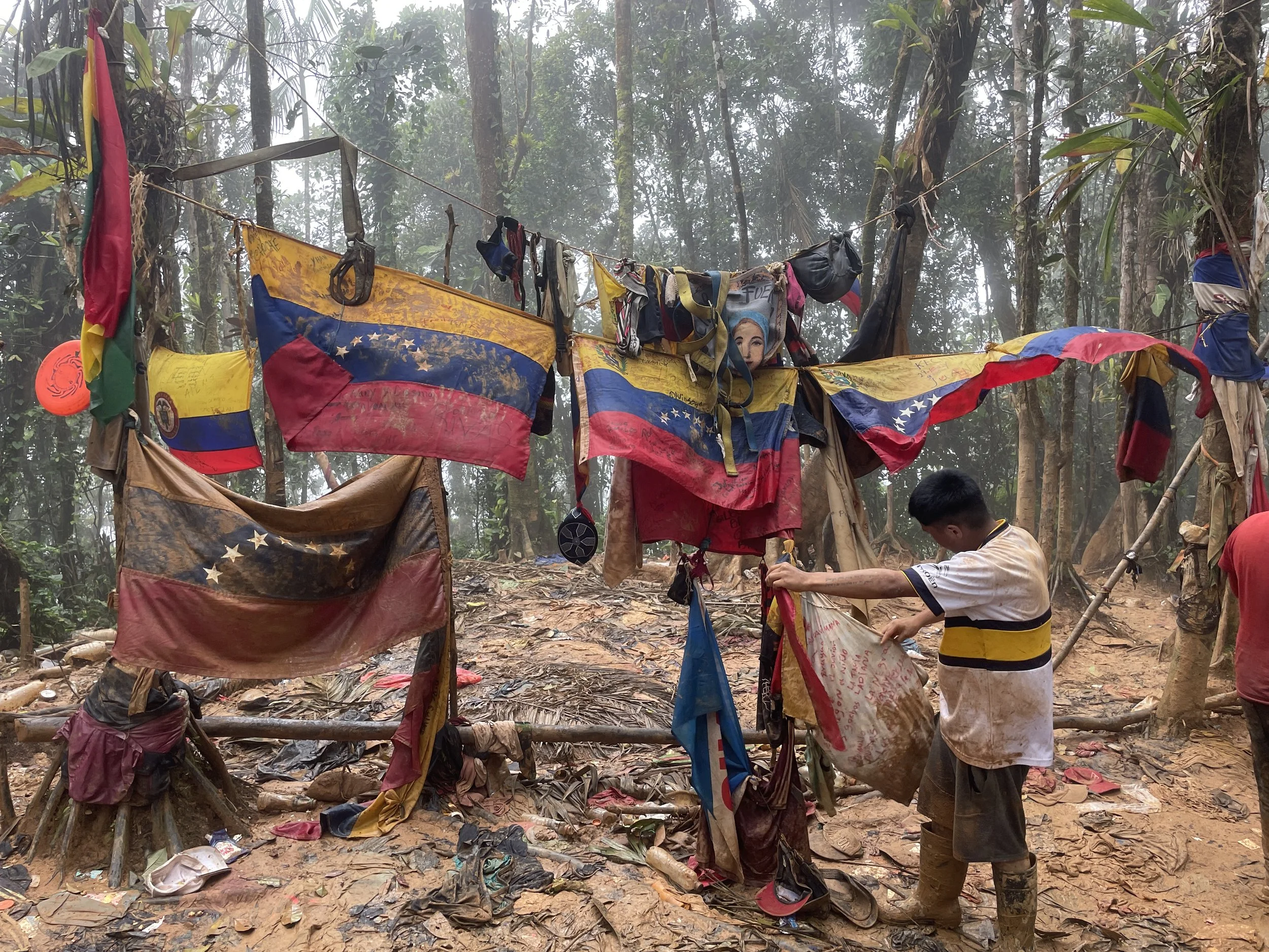 A young man is cleaning a makeshift memorial in a muddy, forested area, with various weathered Venezuelan flags hanging from a wooden structure with rope. The ground is covered with leaves and trash, and the atmosphere appears damp and overcast.