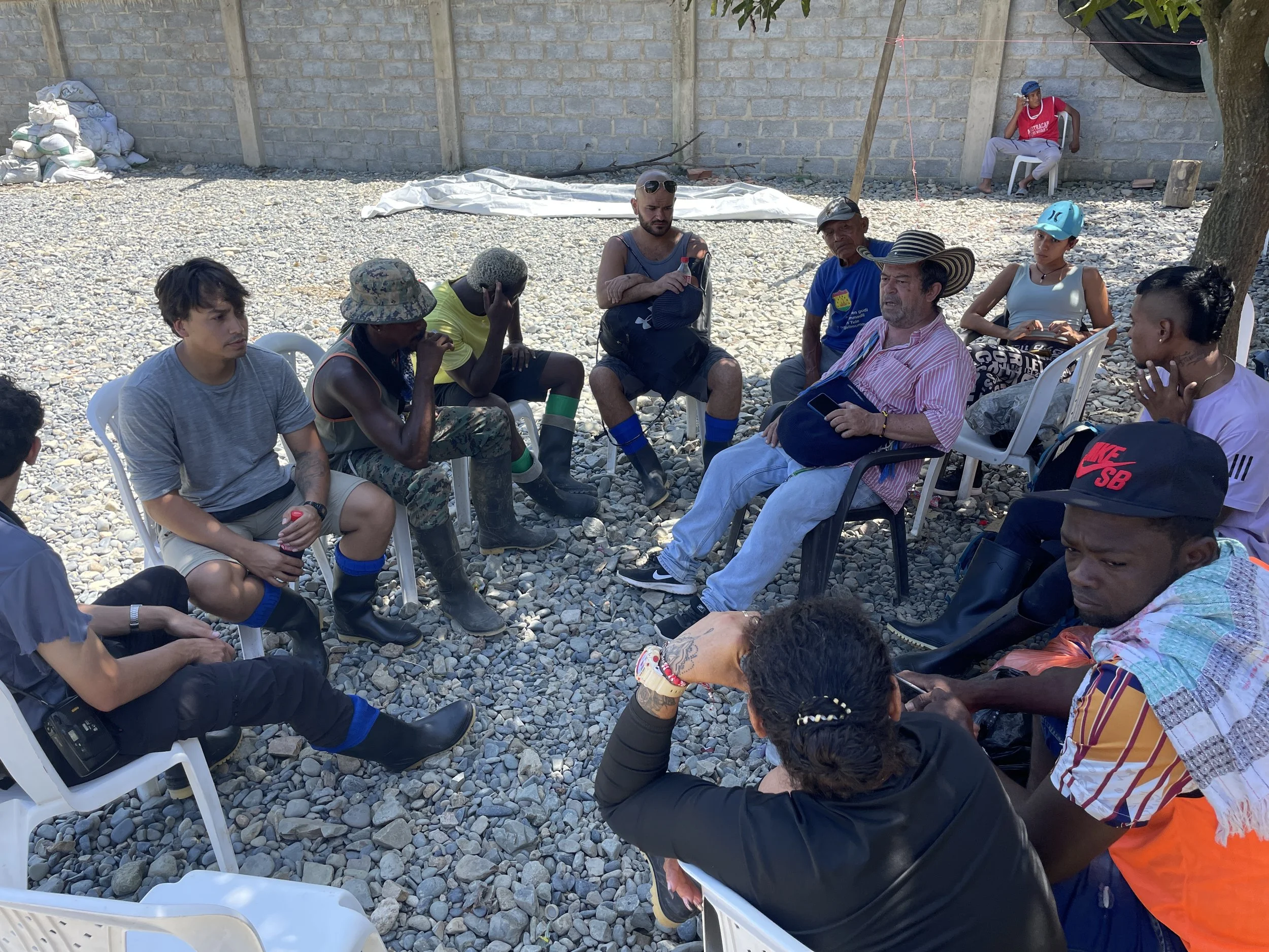 Group of diverse people sitting in a circle outdoors on gravel, engaged in a discussion, with some wearing hats and casual clothing.