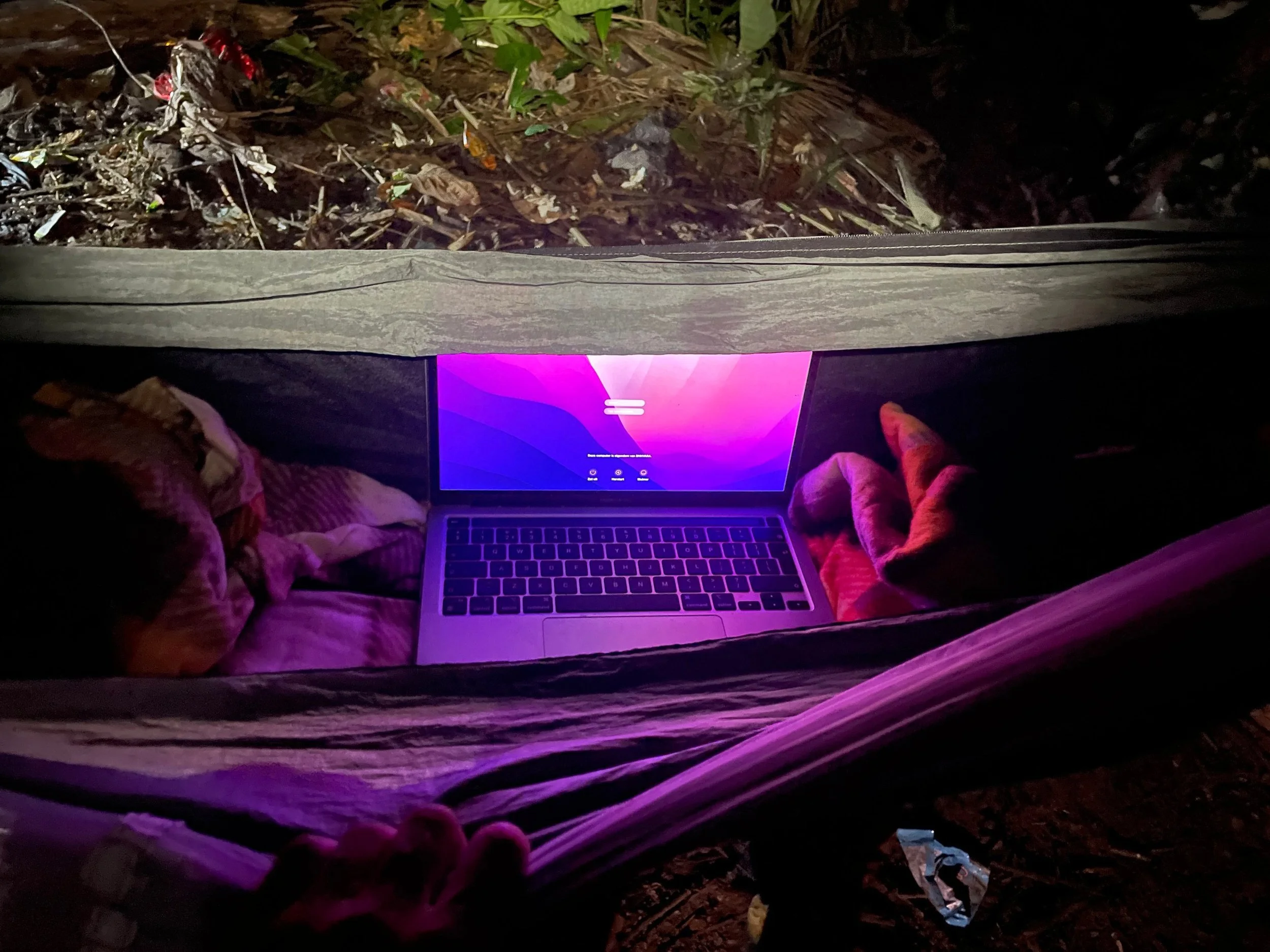 A person lying inside a hammock at night using a laptop. The scene shows the laptop screen glowing with a purple background, and the hammock is suspended above the ground with leaves and dirt visible below.