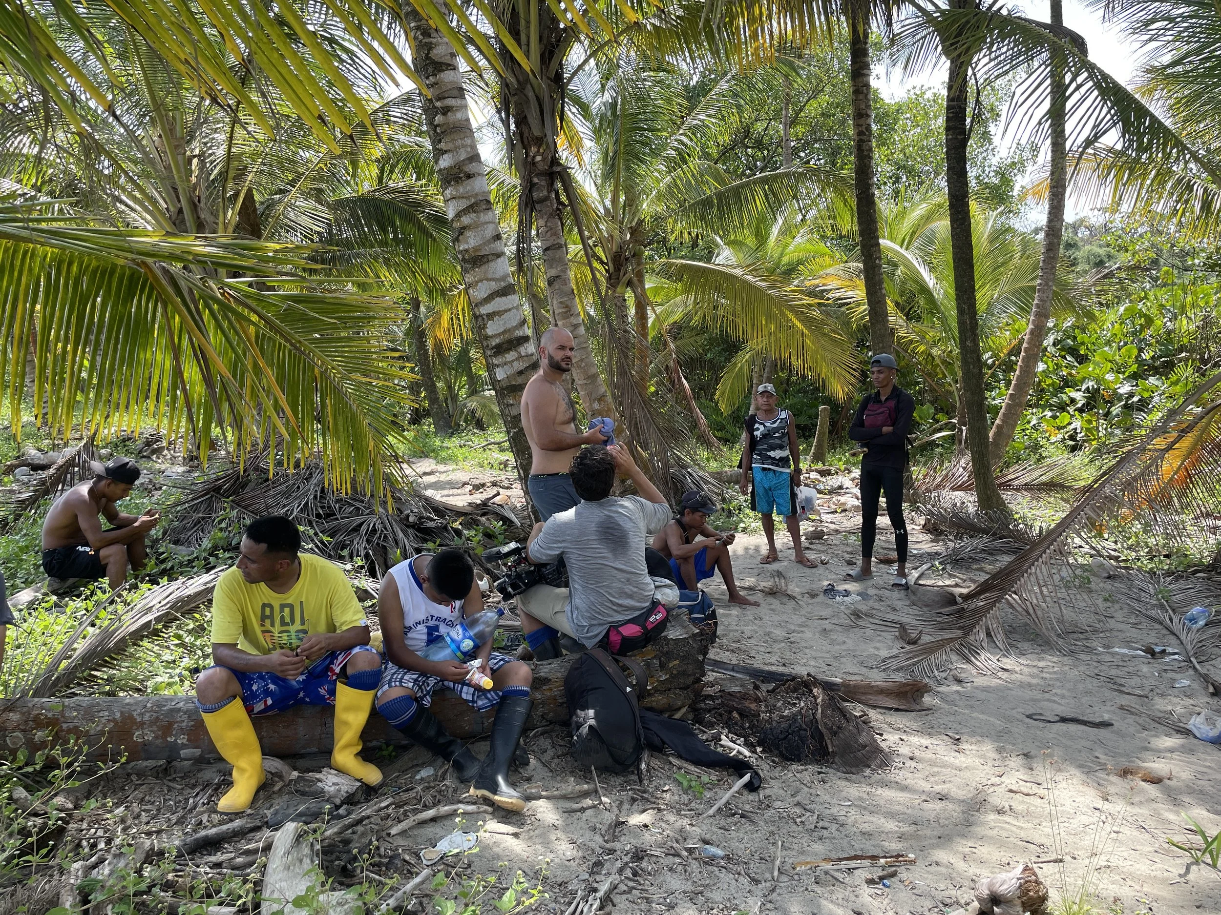 Group of people gathered in a lush tropical forest, some sitting and others standing, under tall palm trees, with a mix of casual clothes and some in colorful rain boots.