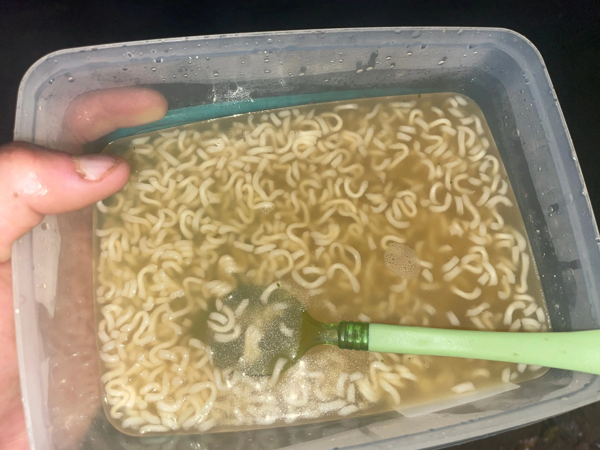 A clear plastic container filled with cooked instant ramen noodles and broth, with a green spoon inside, and a person's hand holding the container.