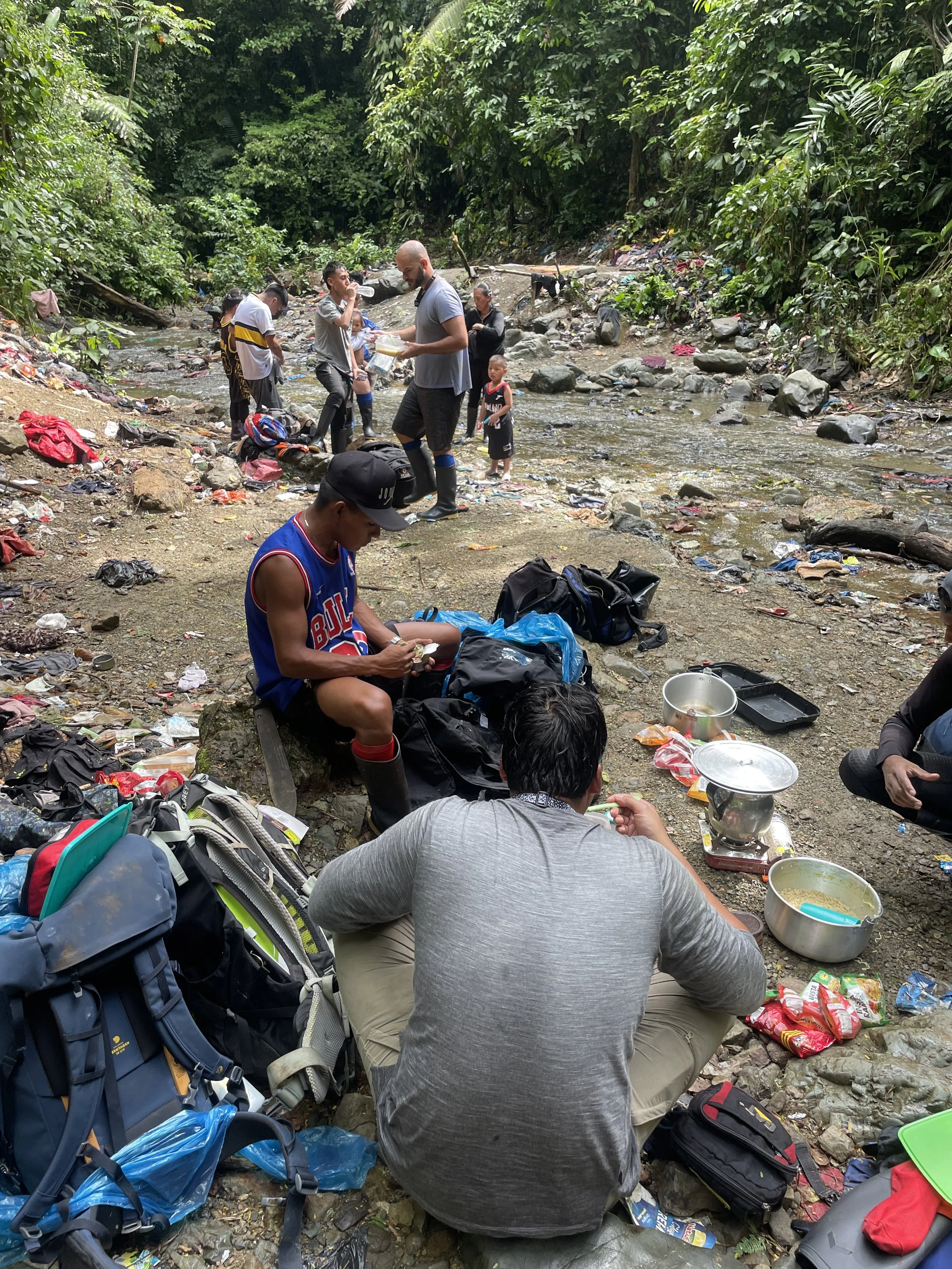 Group of people having a picnic by a small creek in a forested area, with some cooking and eating. There are backpacks and dishes scattered around.