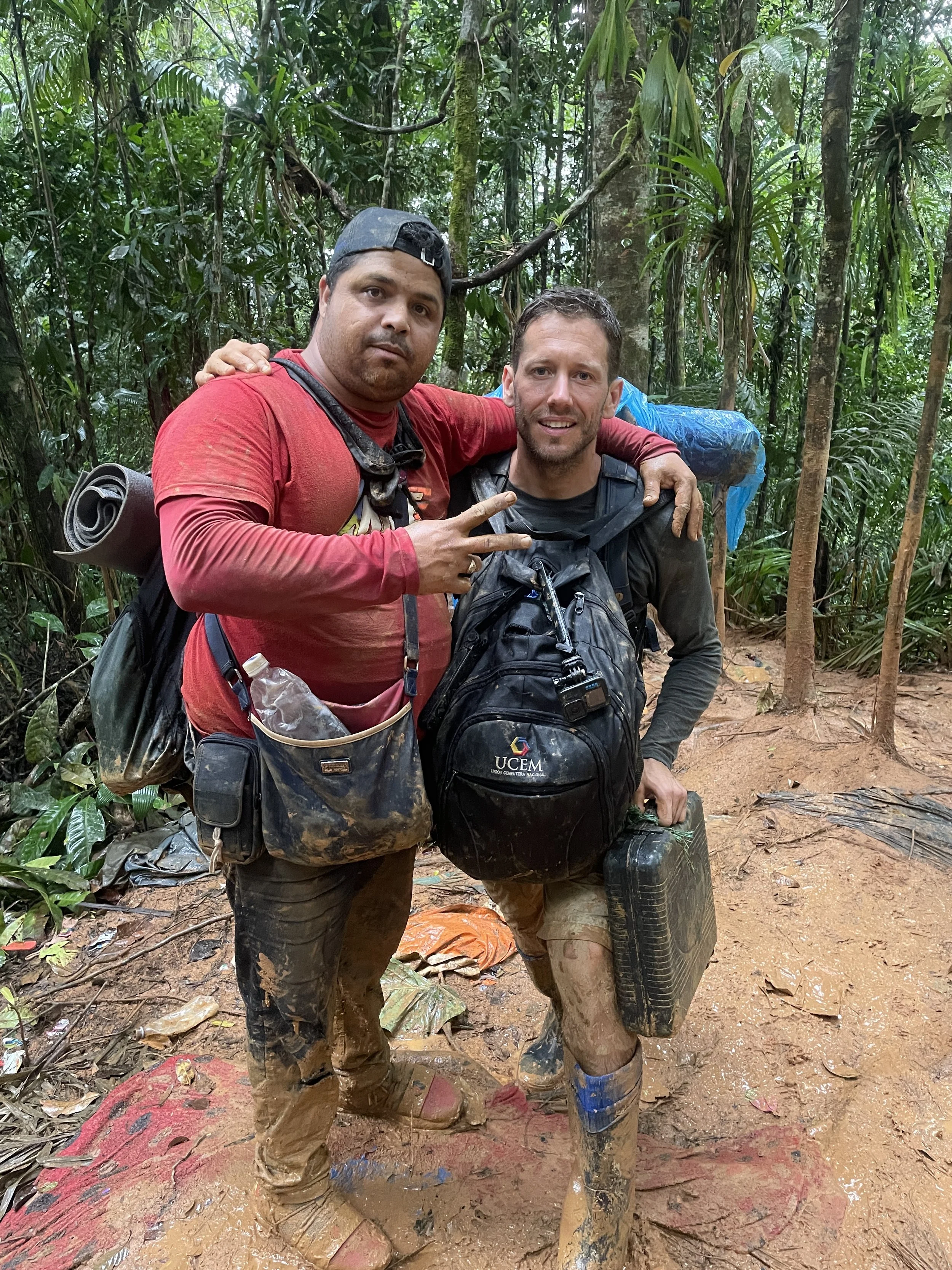 Two men with backpacks and muddy clothes standing in a dense jungle, posing for a photo.