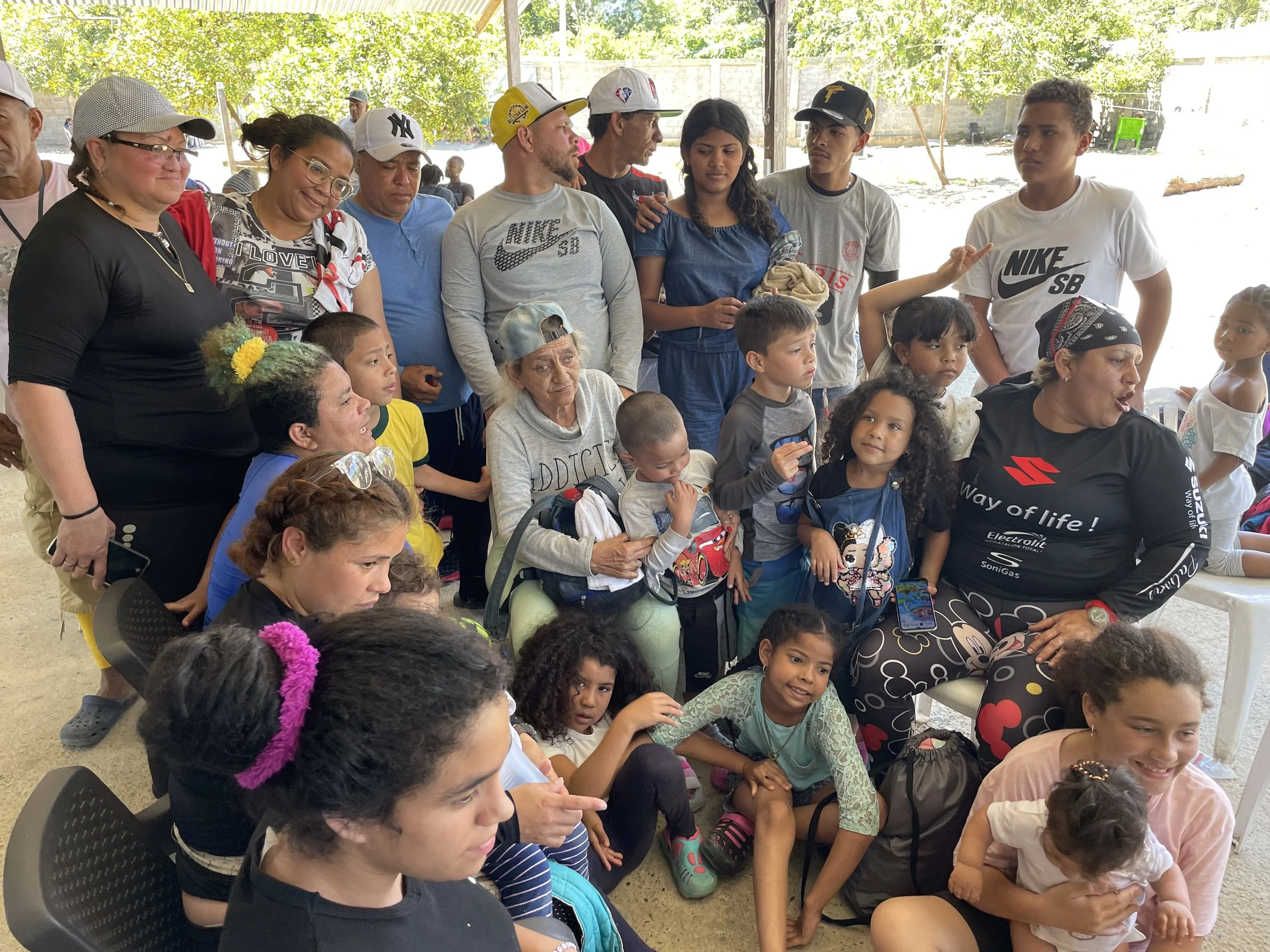 A group of children and adults gathered together indoors, some sitting and some standing, engaged in conversation and activities.