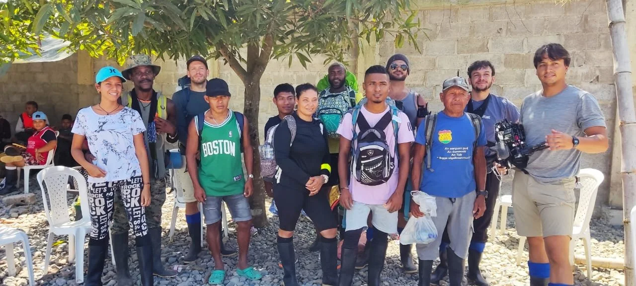 Group of twelve people standing outdoors under a tree, some with backpacks, in front of a stone wall, with chairs and children in the background.