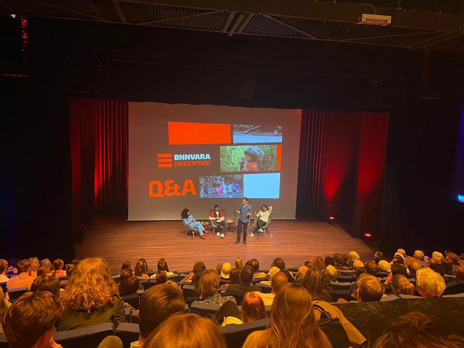 A panel discussion or presentation taking place on a stage in front of an audience in a theater or auditorium. The large screen behind displays the words "BNNVARA NAGESPREK" and "Q&A" along with several images, with three people seated on the stage a