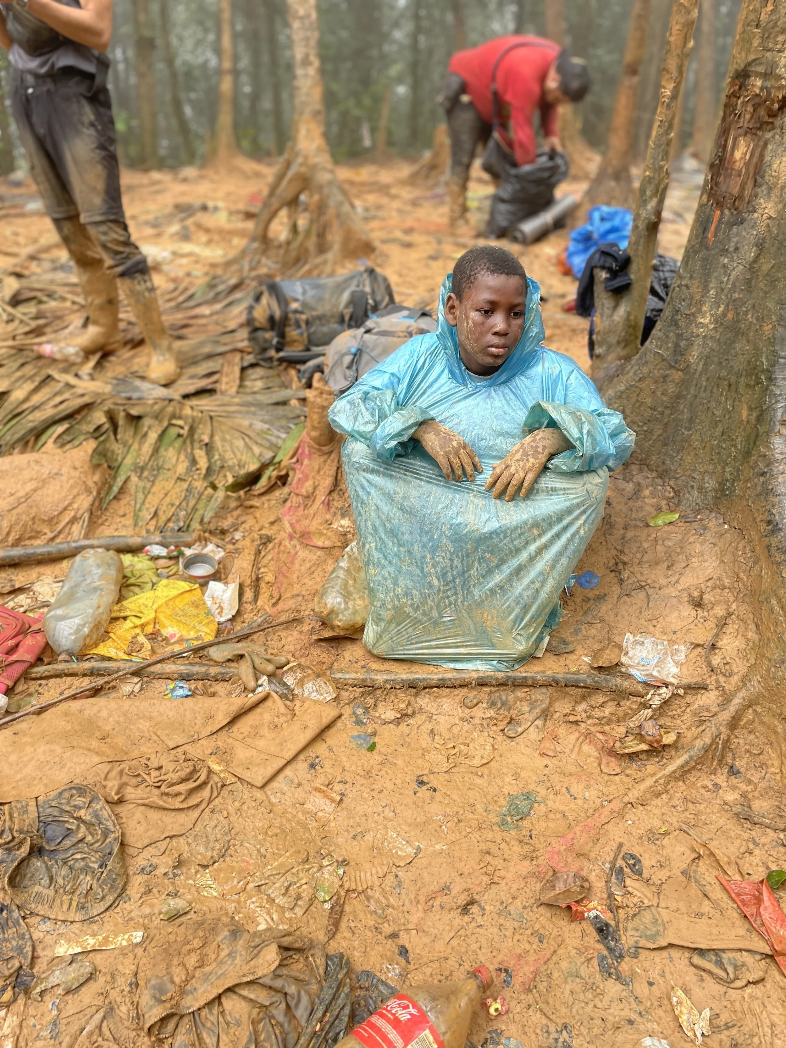 Young boy wearing a blue raincoat and gloves in a muddy forest, surrounded by discarded trash, with other people in the background also collecting waste.