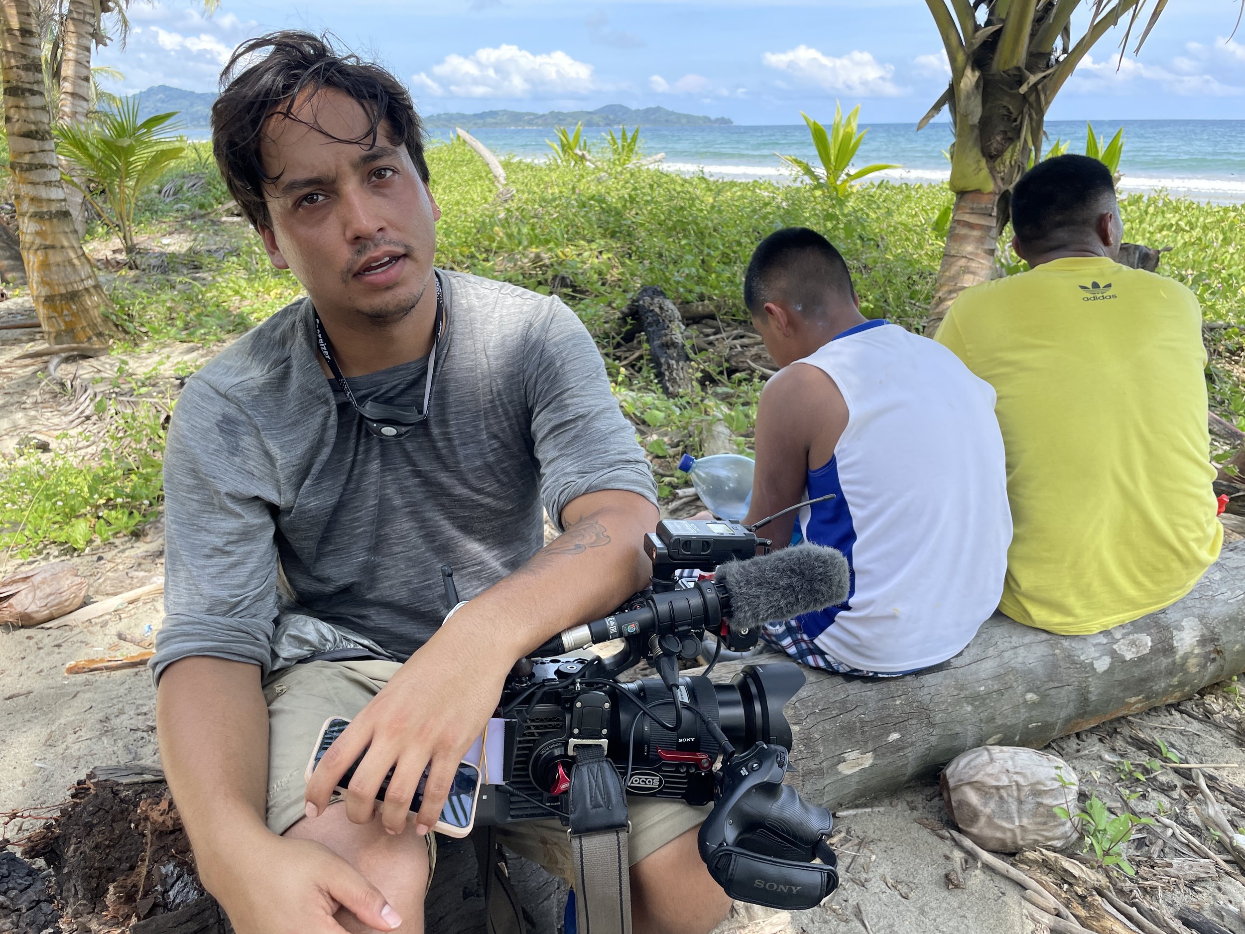 A young man, with a camera and microphone, sitting on a log on a sandy beach with lush greenery, palm trees, and the ocean in the background, while two other men sit on the log with their backs to the camera.