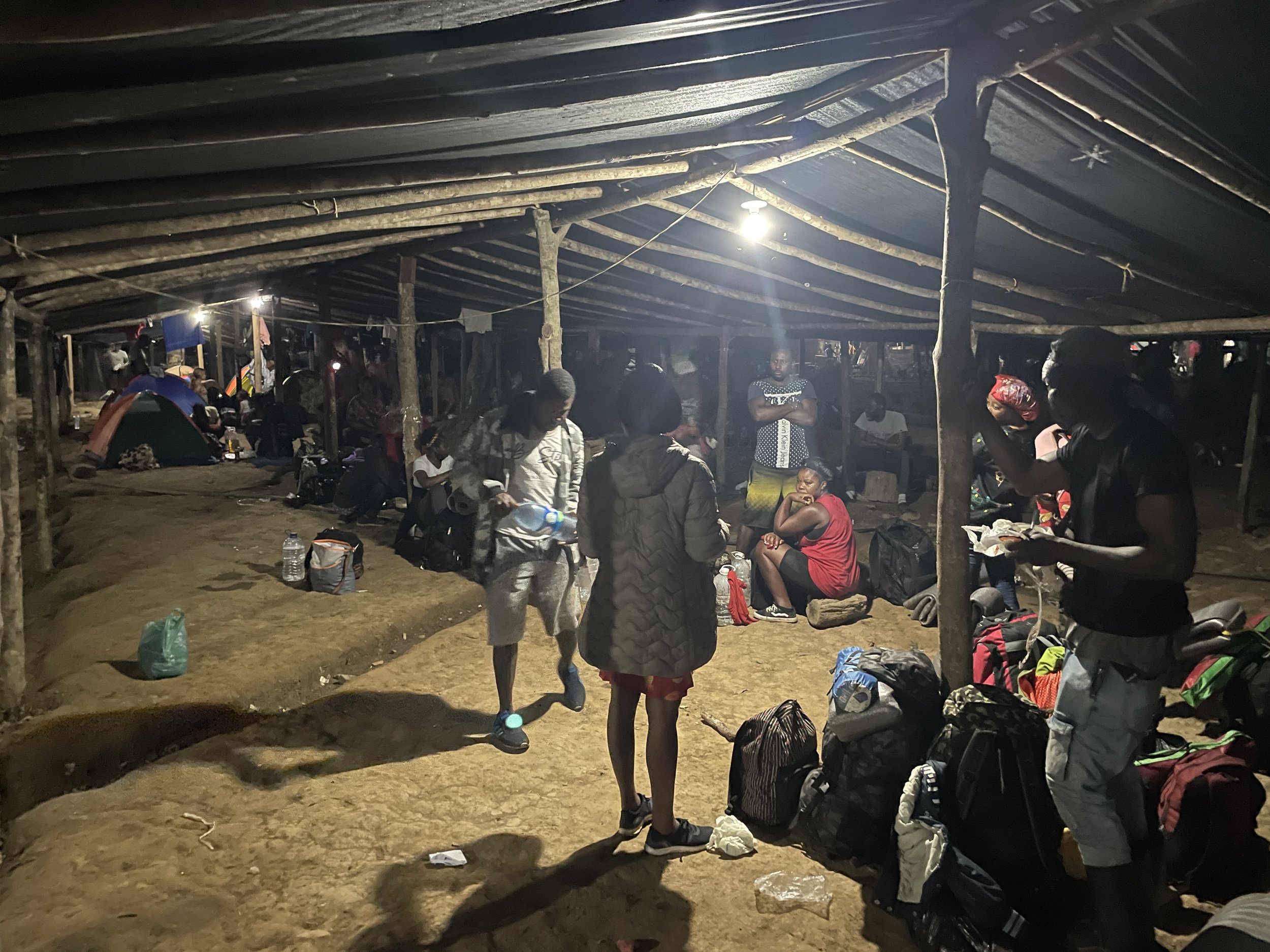 Nighttime scene inside a makeshift shelter with people sitting and standing, surrounded by backpacks and belongings, illuminated by a single overhead light.