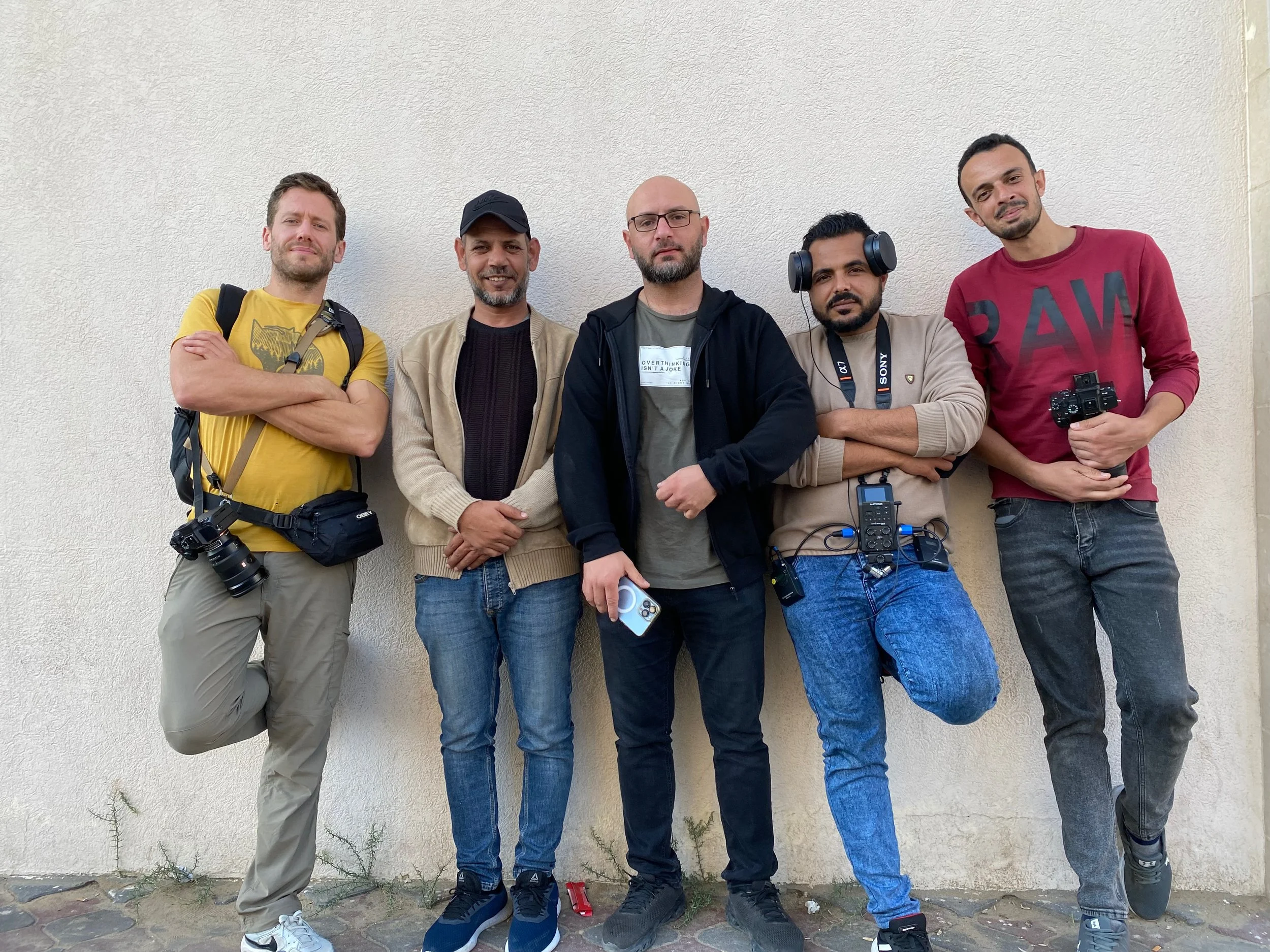 Group of five men posing outdoors against a plain wall, some holding cameras and equipment.