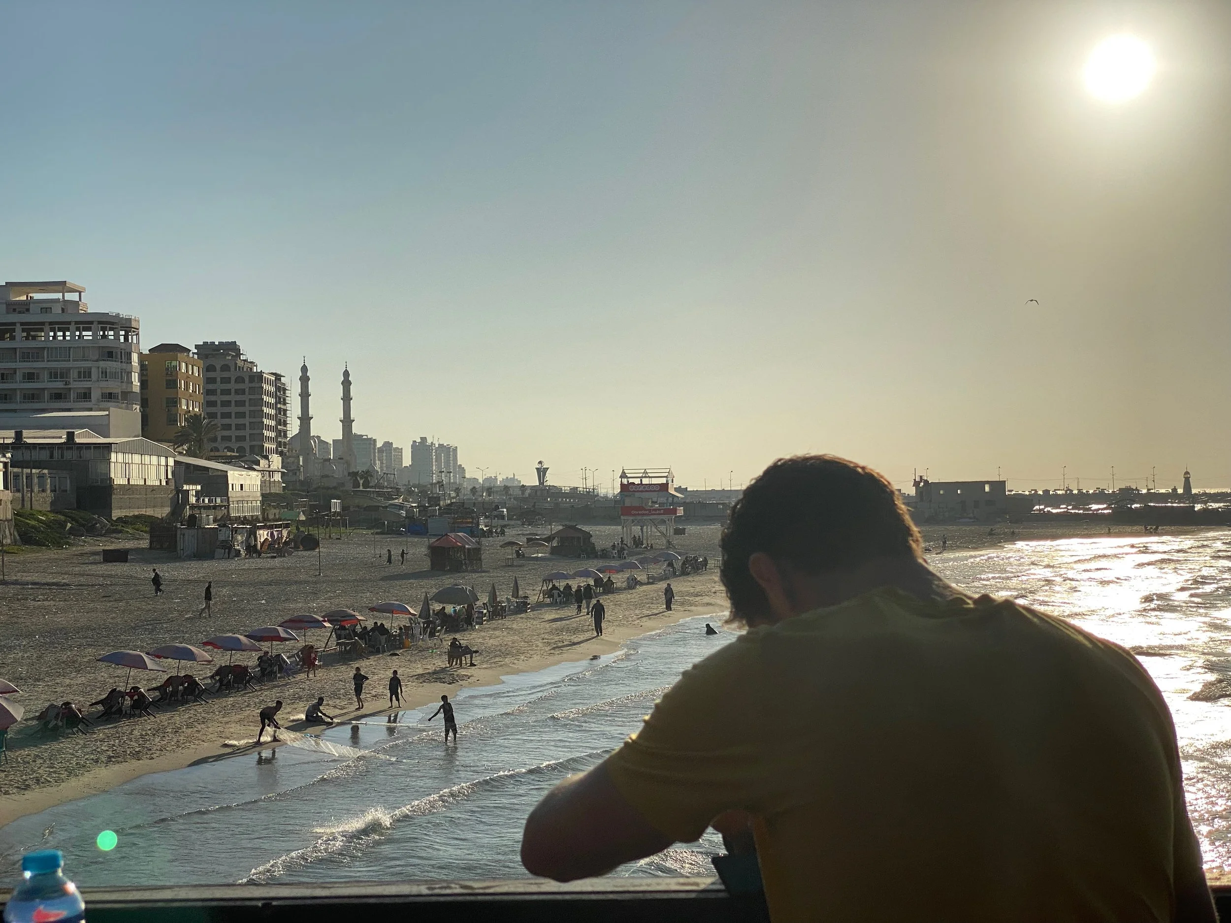 Person sitting by a beachside with a city skyline and mosques in the background, sun setting, umbrellas on the beach, people walking, and the sea.