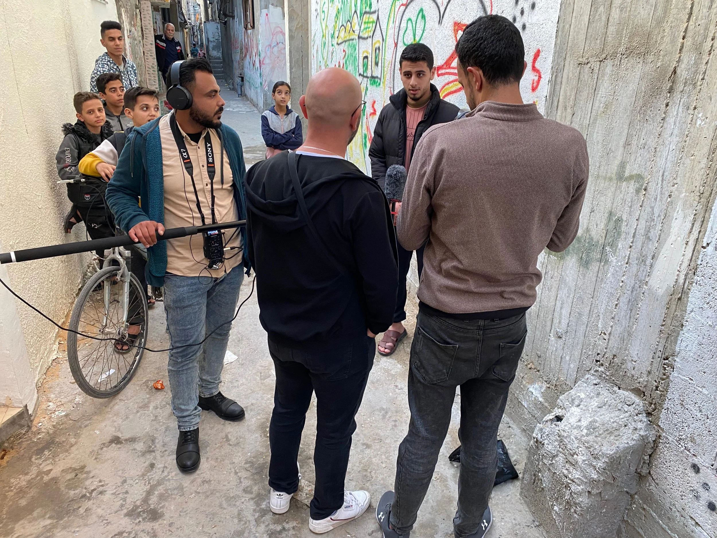 A group of people, including a man with headphones and camera equipment, are gathered in a narrow alleyway with graffiti-covered walls, engaged in a conversation or interview.