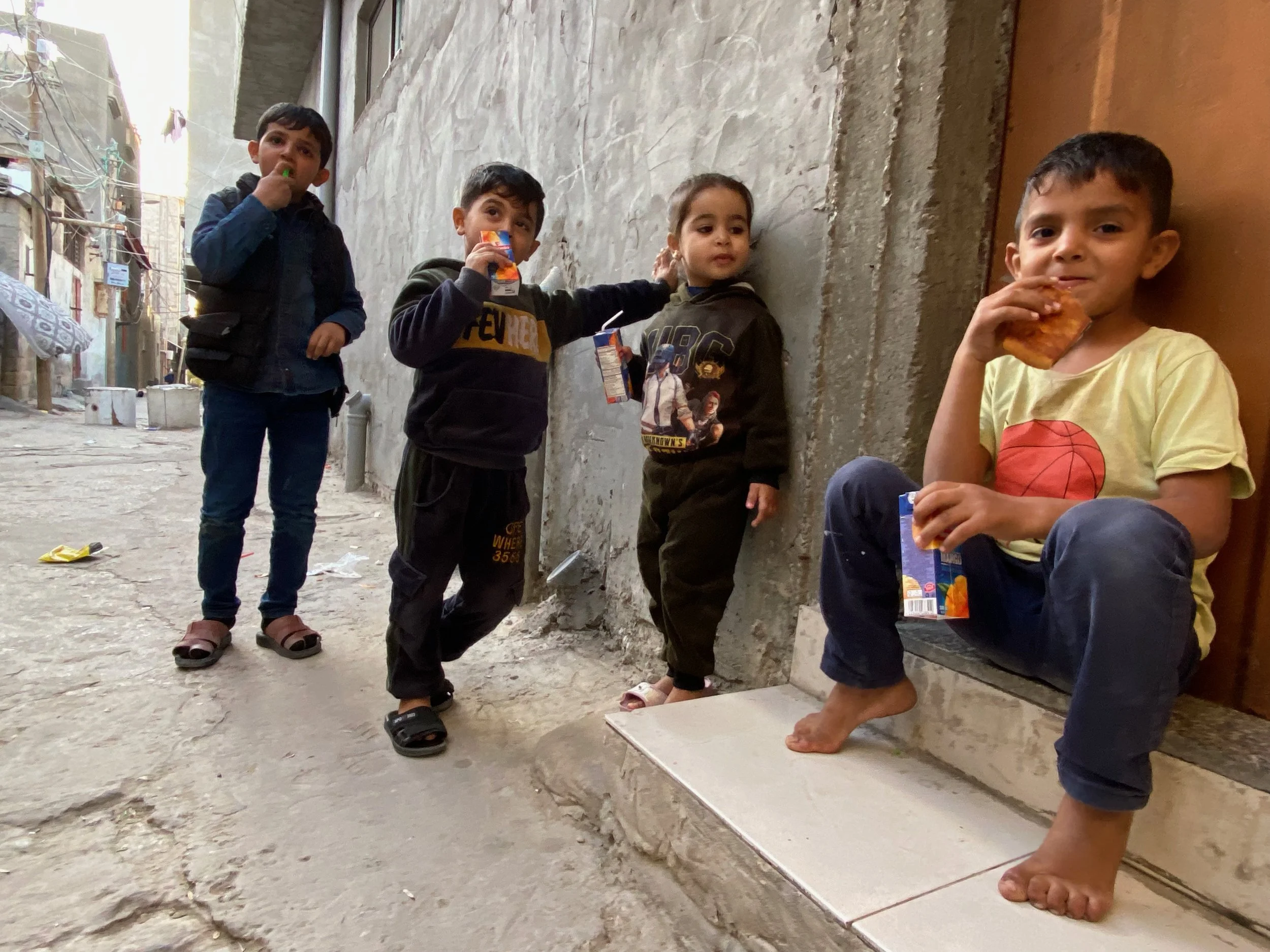 Four young boys standing and sitting on a sidewalk against a weathered wall, eating snacks and holding juice boxes in an alleyway.