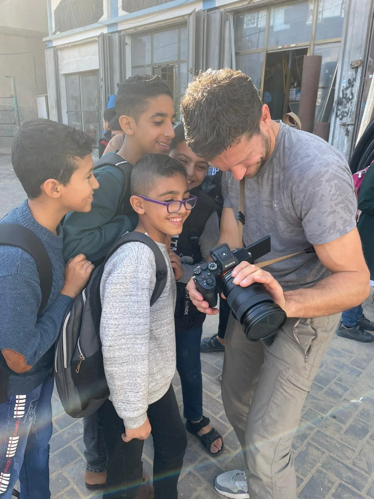 A man showing a camera to a group of children, all smiling and gathered around him outdoors.
