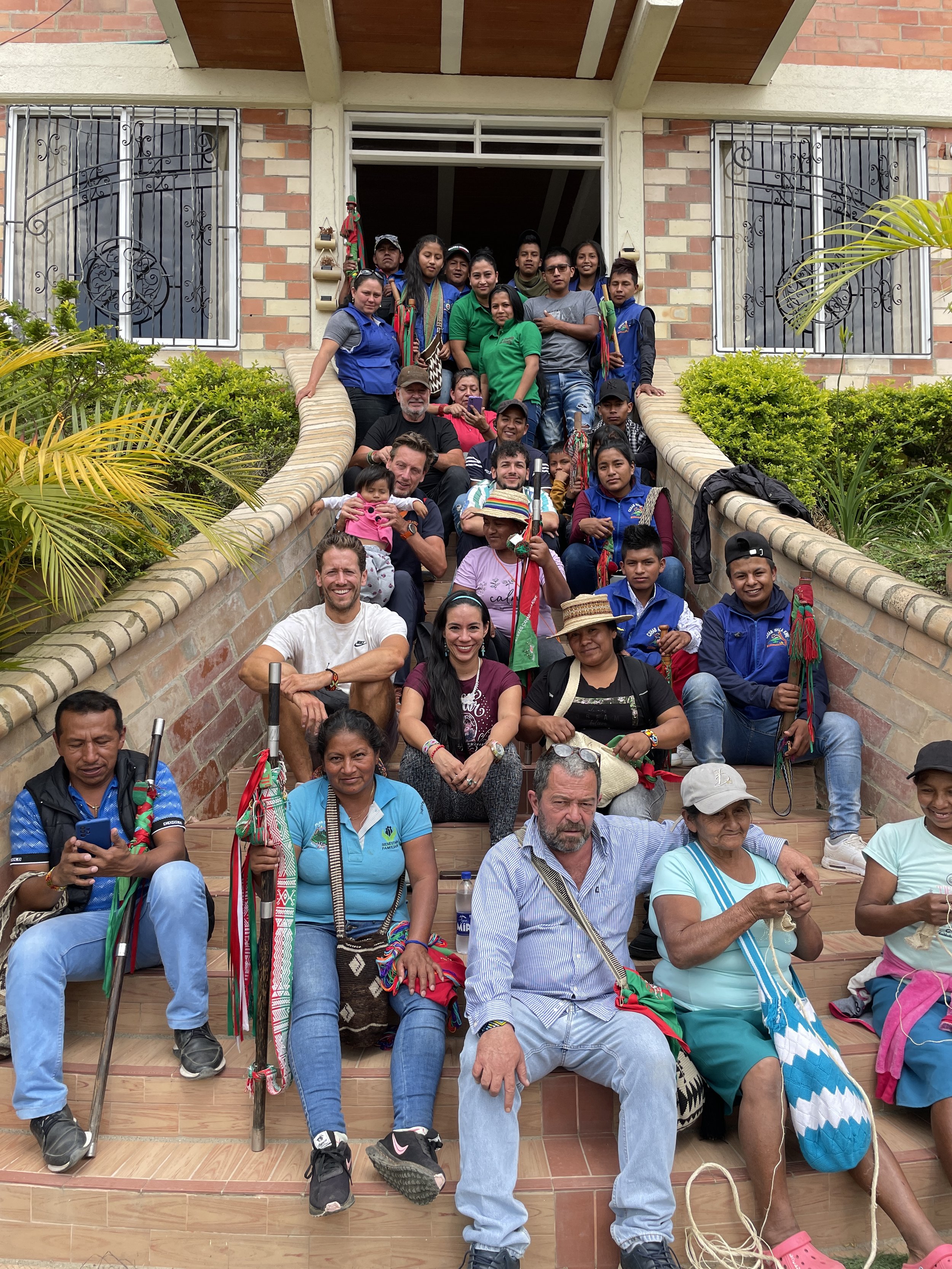 Group of people gathered on the steps of a building, some holding traditional items, with diverse ages and ethnic backgrounds, in front of brick and concrete house with decorative window bars and greenery.