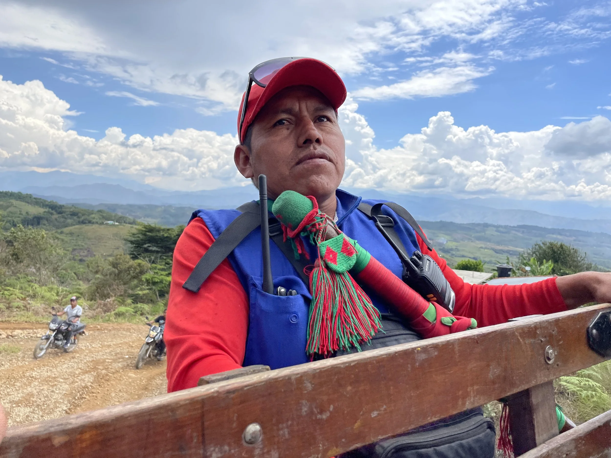 A man in a red and blue outdoor outfit with a red cap and sunglasses on his head sits on a wooden bench in a rural landscape with mountains and cloudy sky in the background.