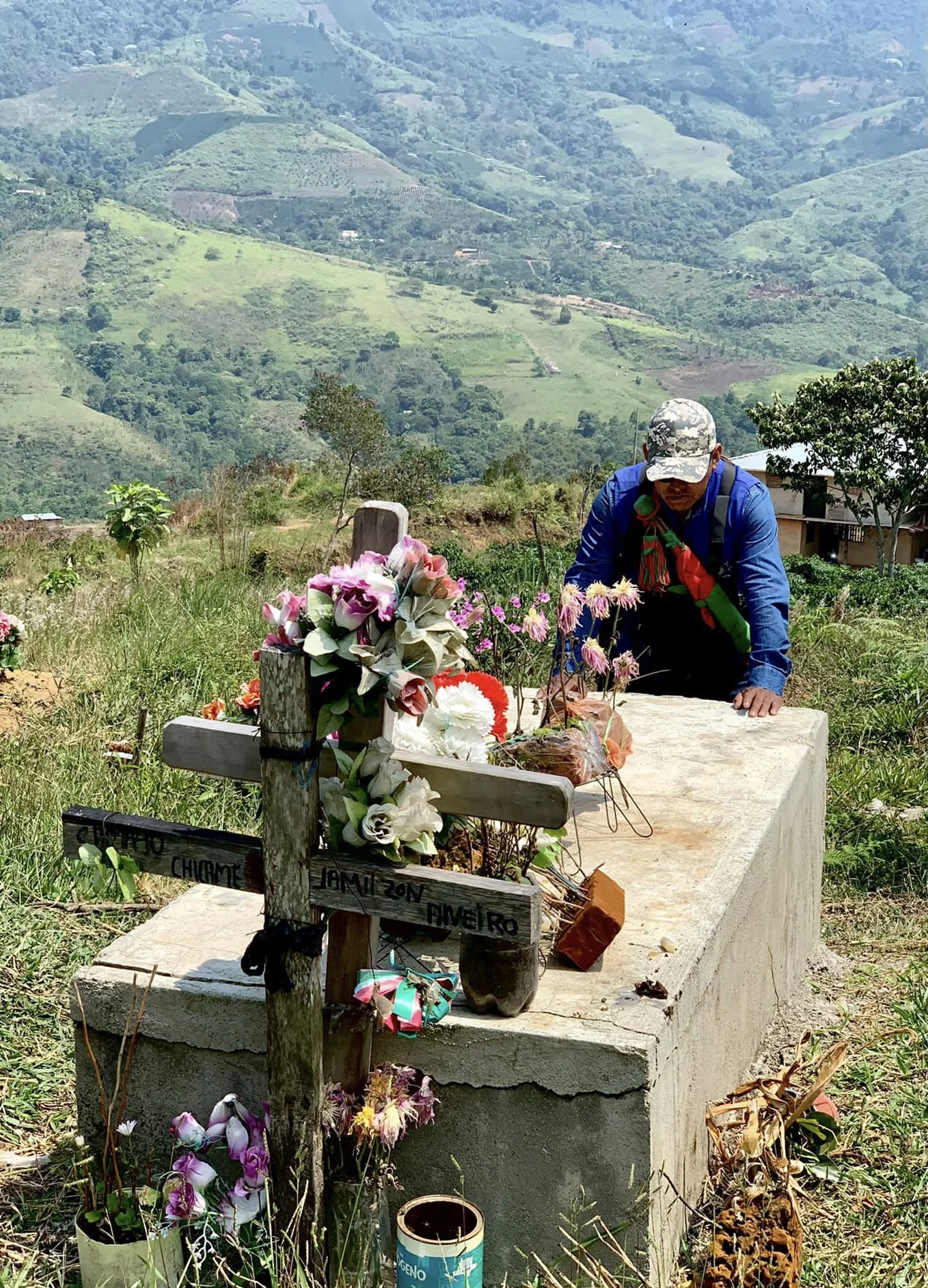 A person in a blue shirt, camouflage cap, and scarf kneels at a grave with a wooden cross, flowers, and heartfelt items in a grassy field with lush green hills in the background.