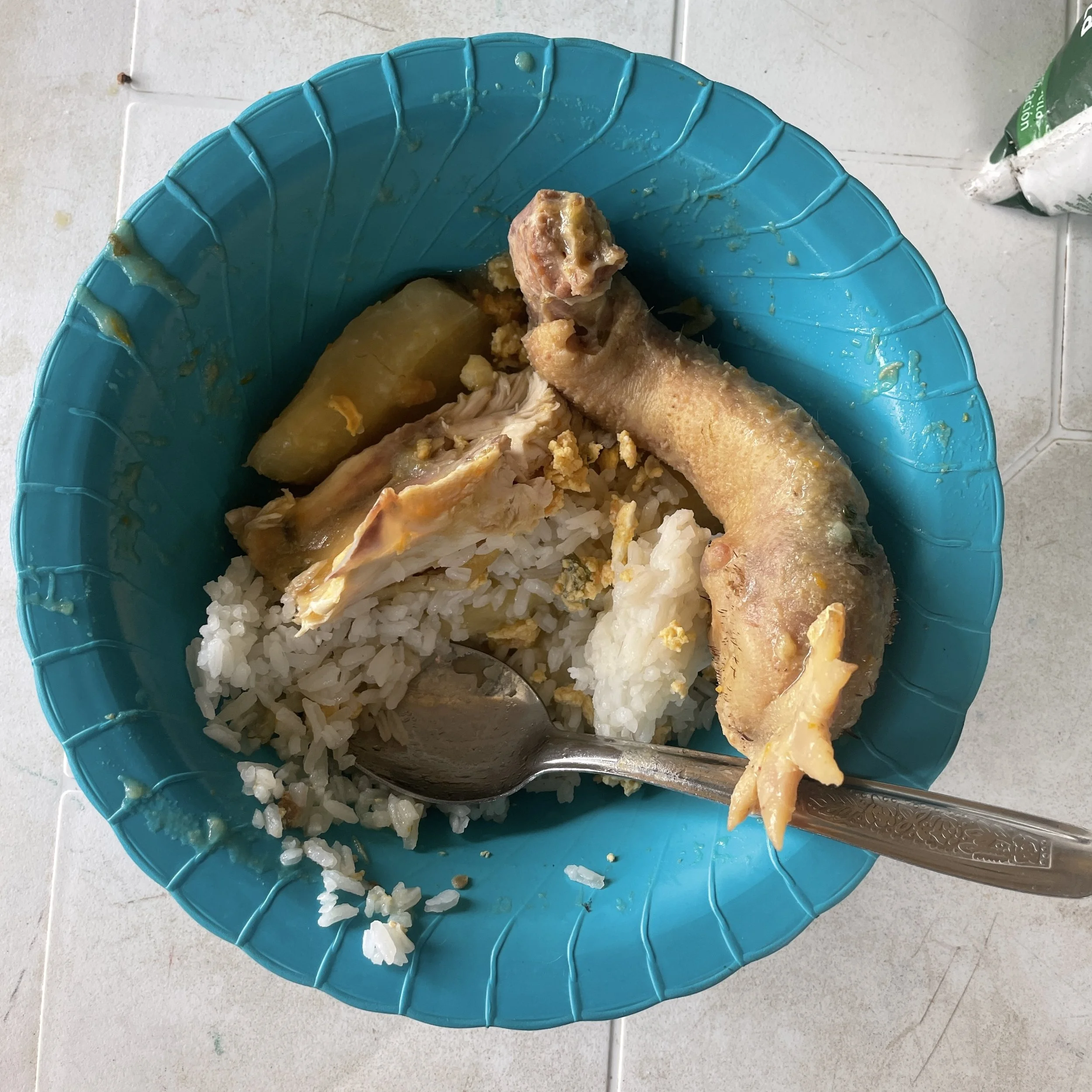 A blue plastic bowl contains rice, a chicken drumstick, a potato, and some sauce or gravy, placed on a white tiled surface.