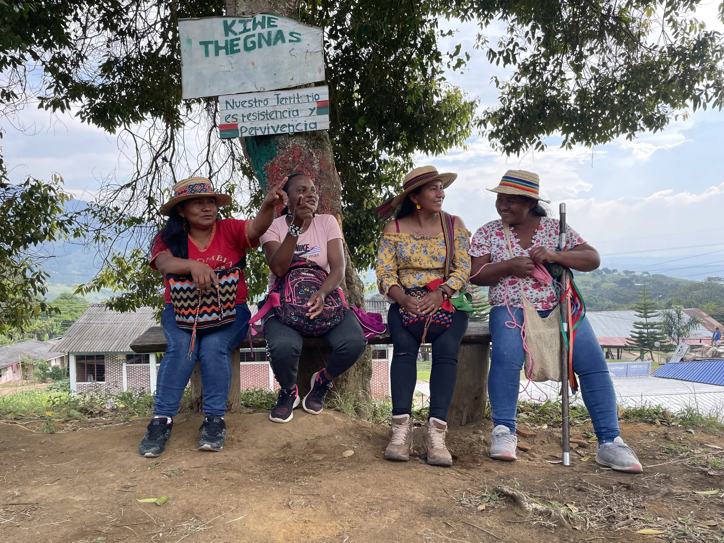 Four women sitting on a bench in front of a tree, with village houses and green hills in the background. They are wearing colorful clothing and straw hats, and one is knitting with pink yarn. A sign on the tree reads 'KIWE THEGNAS' and 'Nuestro Terri