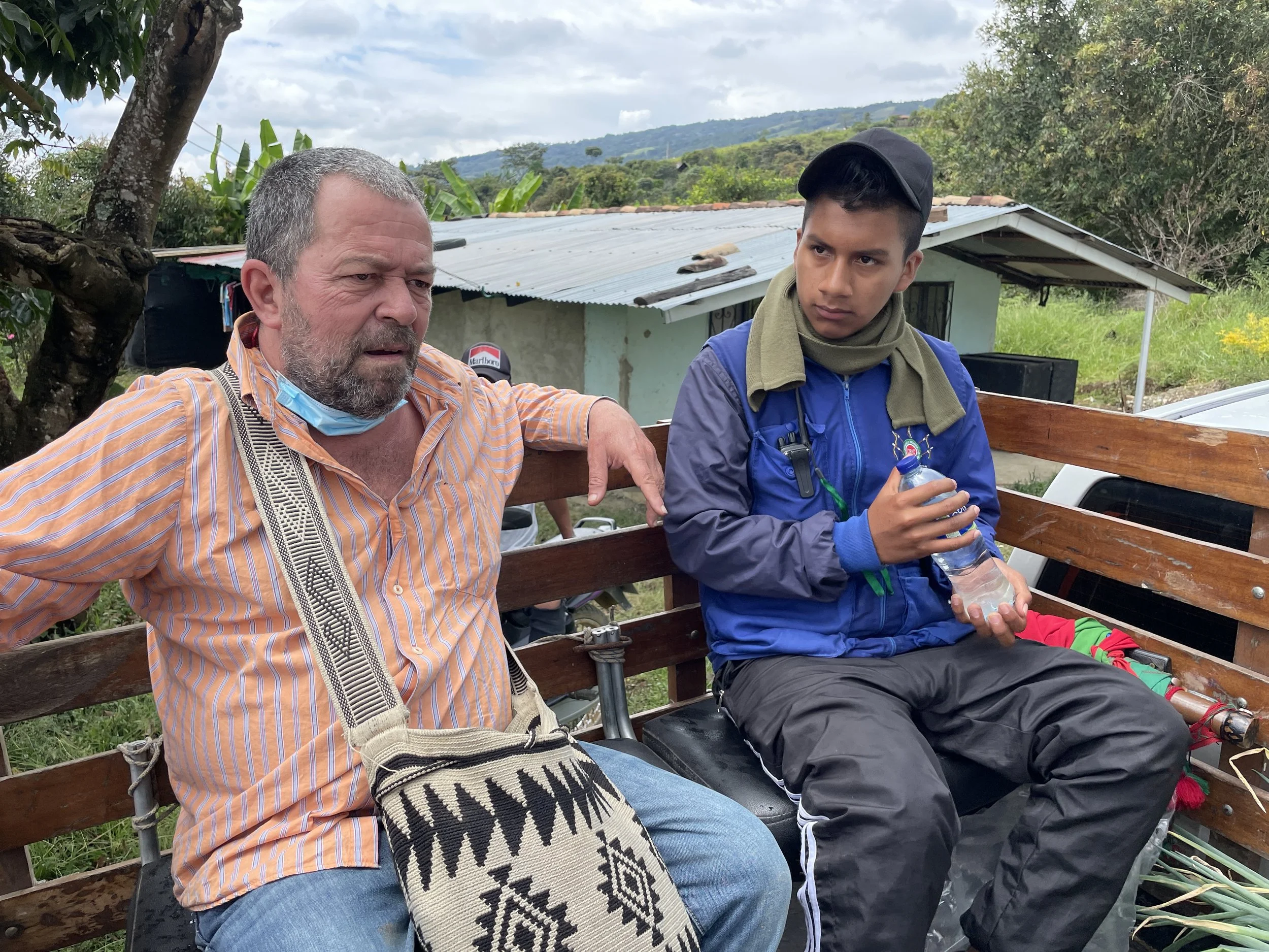 Two men sitting on a wooden bench outdoors, one older with a beard wearing an orange striped shirt, and a younger man in a blue jacket holding a water bottle, with hills and a house in the background.