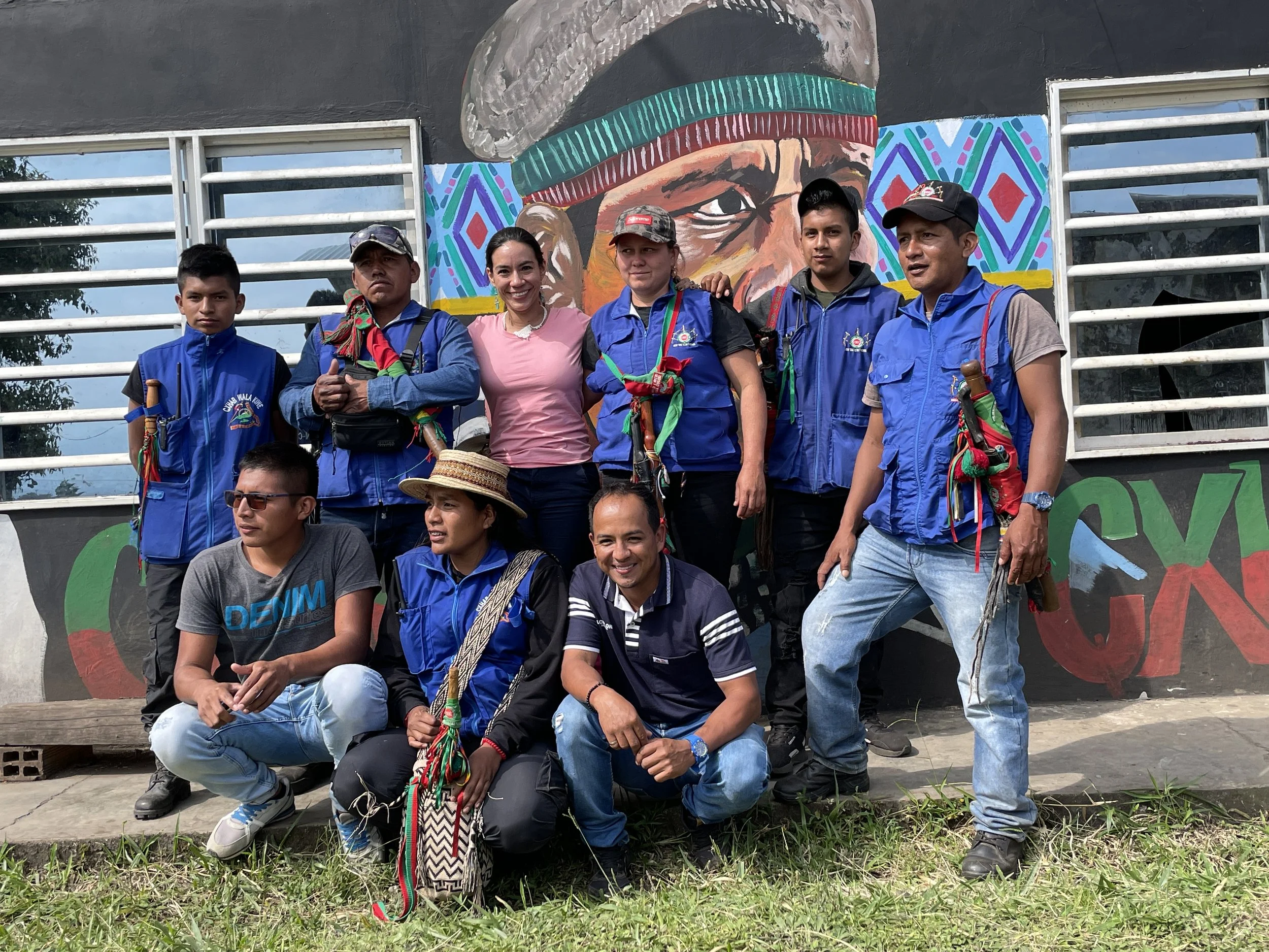 Group of nine people, some wearing blue vests, posing outdoors in front of a colorful mural mural. The mural features a large portrait of a person wearing a head covering with a geometric background. Some members of the group are kneeling and others 