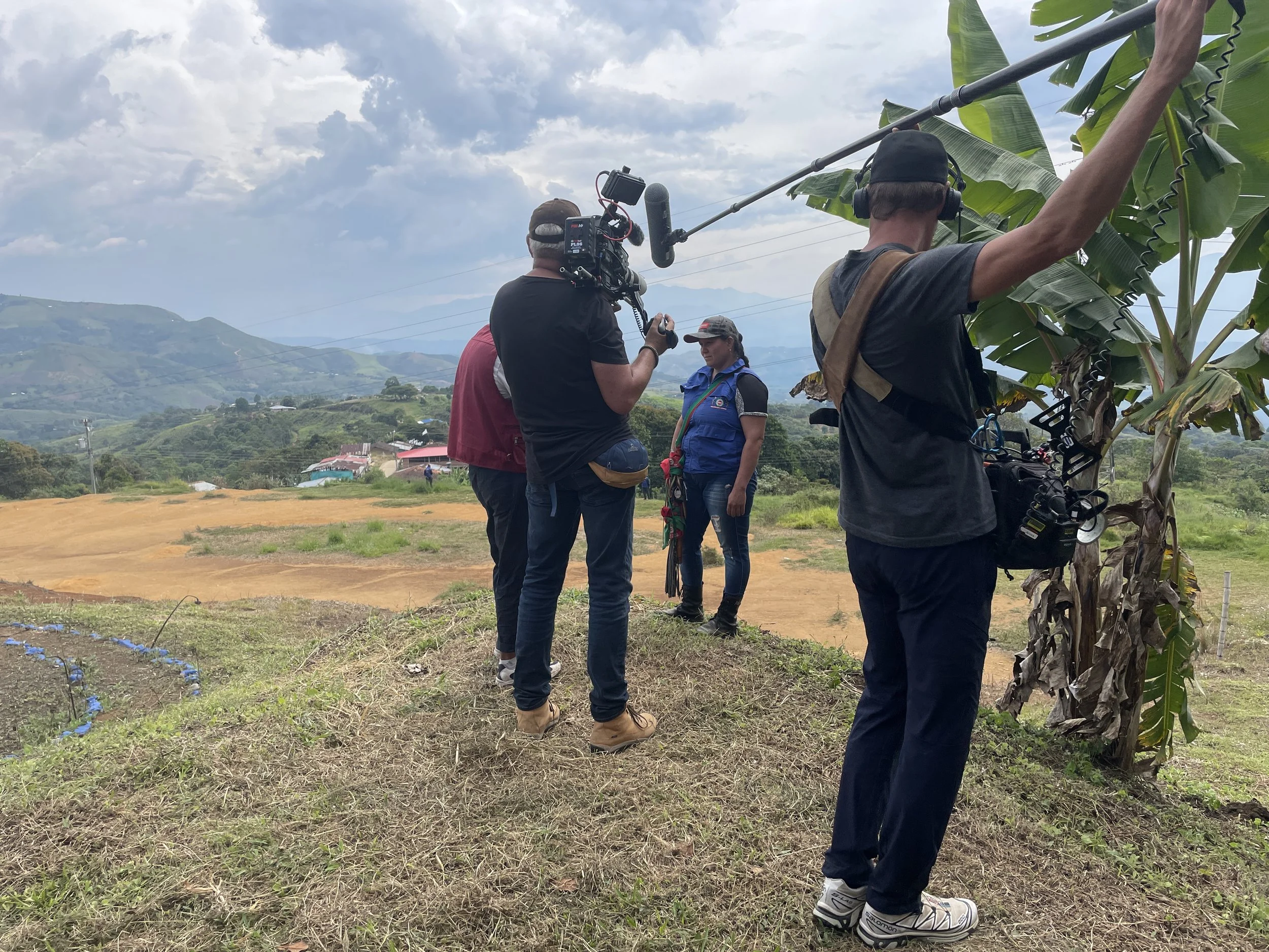 Filming crew on location in a rural area with rolling hills, using cameras, microphones, and headphones, as a woman in a vest stands nearby, with a banana tree in the foreground.