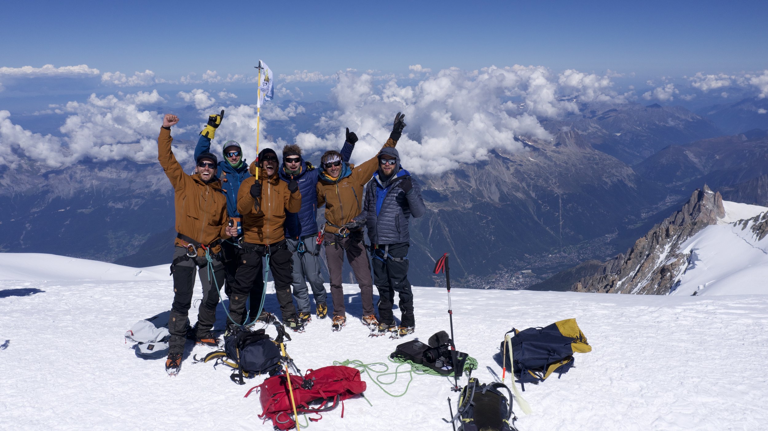 Six climbers celebrating on a snowy mountain peak with ice axes and backpacks, with a mountain range and clouds in the background.