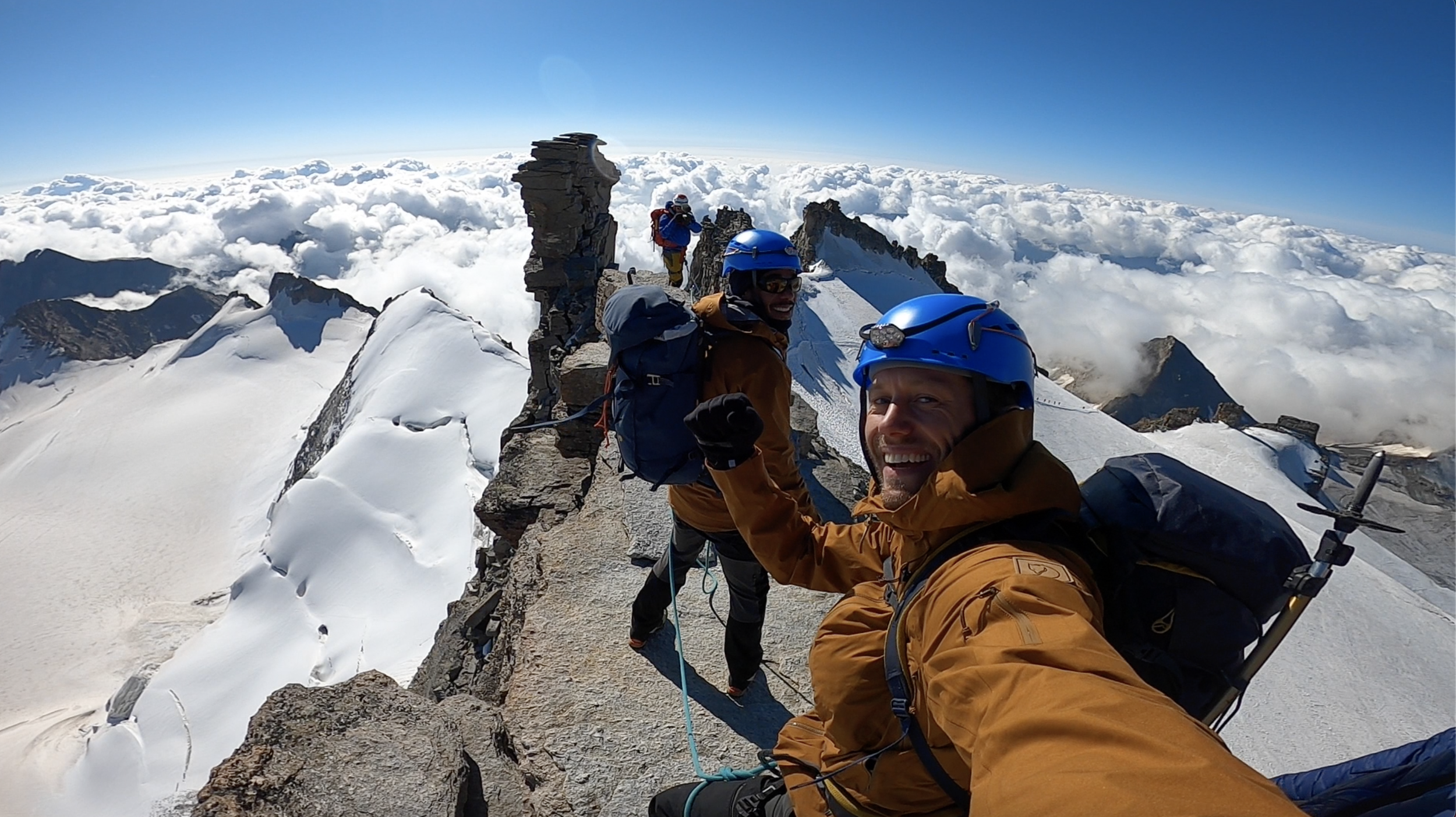 Three climbers wearing blue helmets and brown jackets taking a selfie on a snowy mountain summit with a view of clouds and mountain peaks in the background.