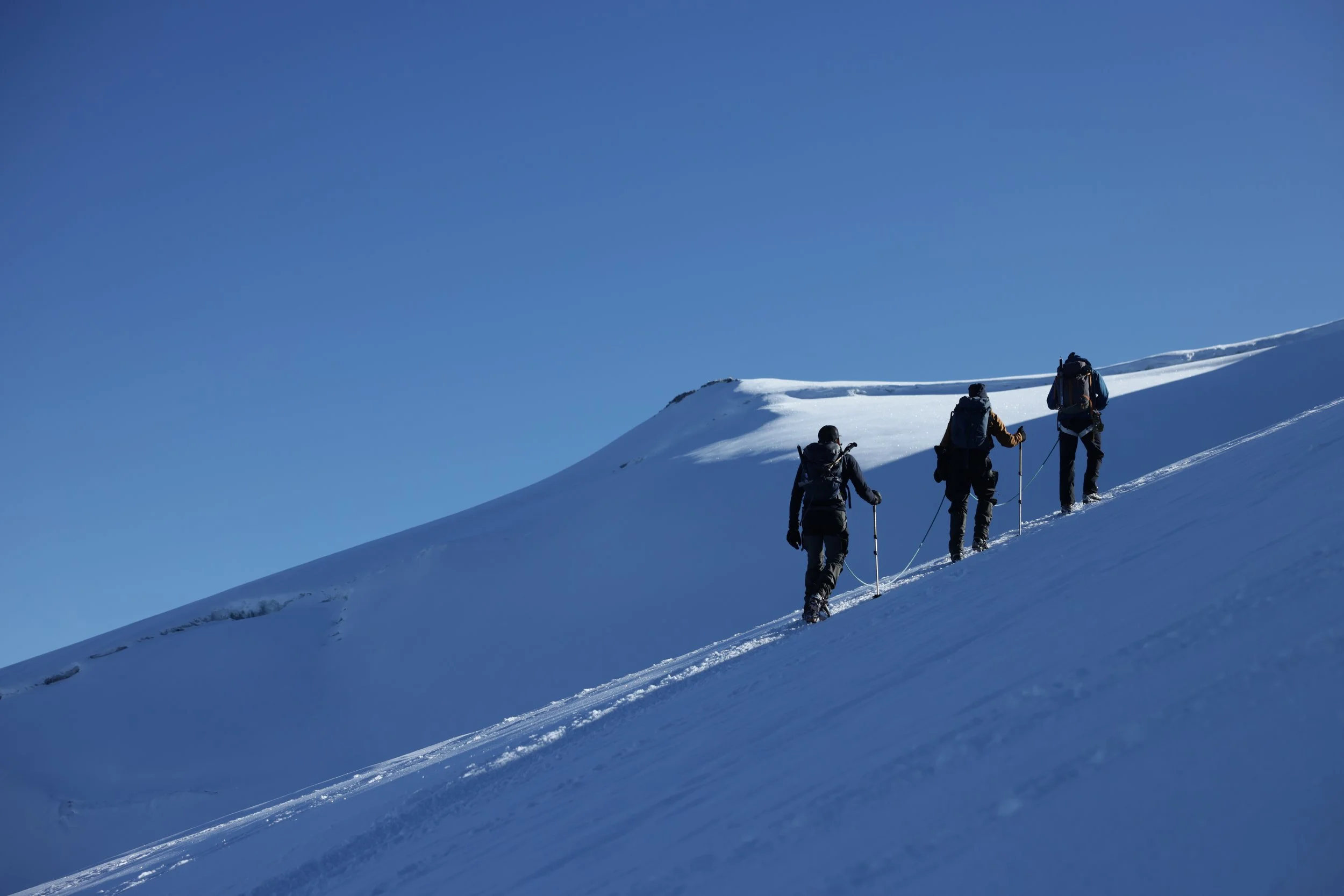 Three hikers trekking up a snow-covered mountain slope under a clear blue sky.
