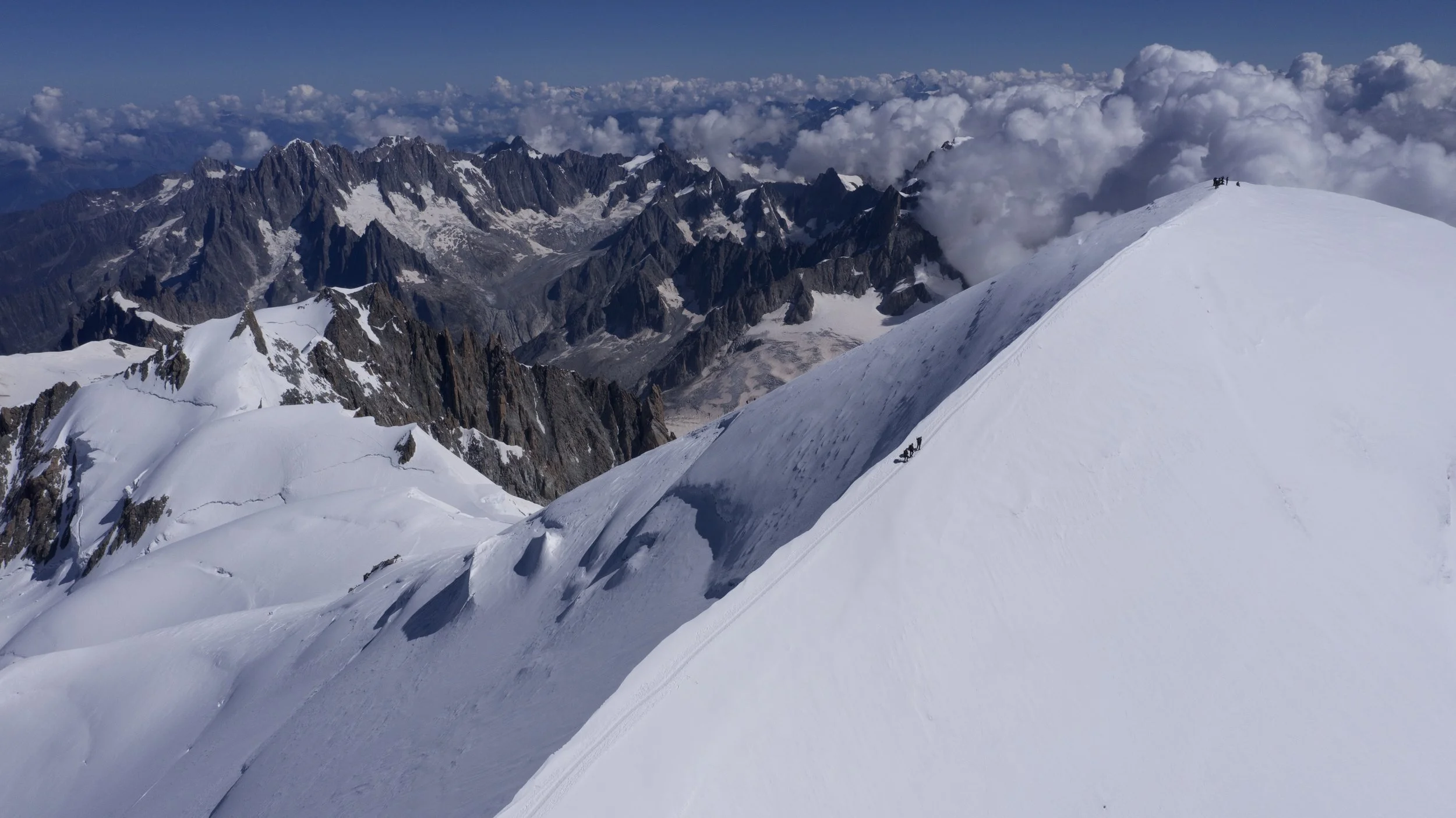 Snow-covered mountain peak with climbers ascending along a trail, high above clouds and rugged mountain range in the distance.