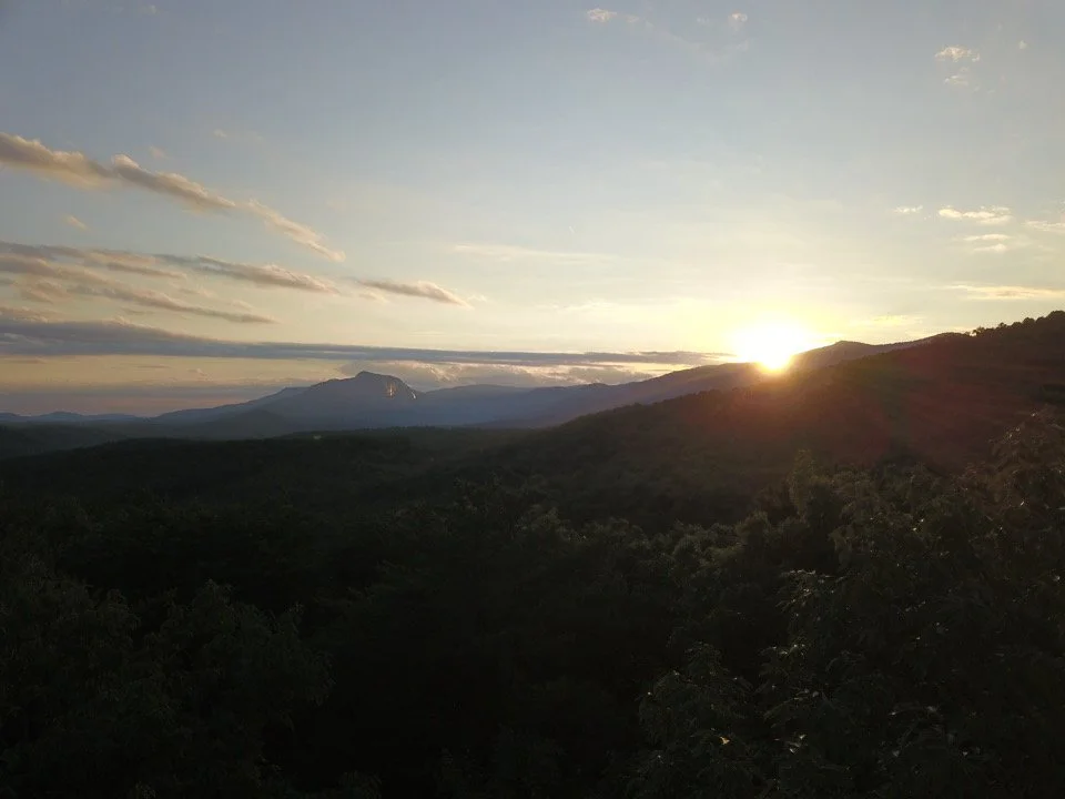 Sunset over a mountain range with trees in the foreground and clouds in the sky.