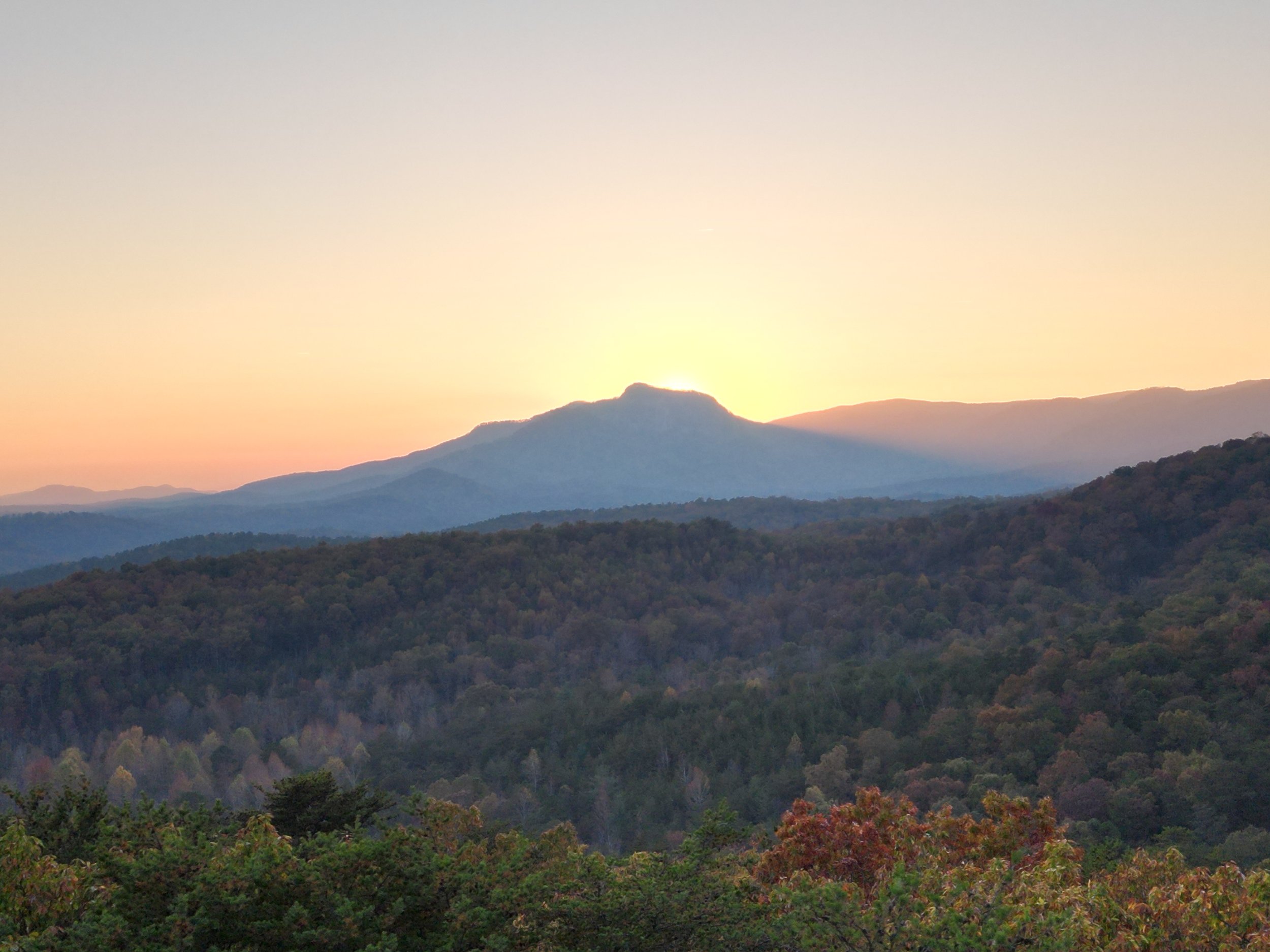 Sunrise over a mountain range with rolling hills and a forested area in the foreground.