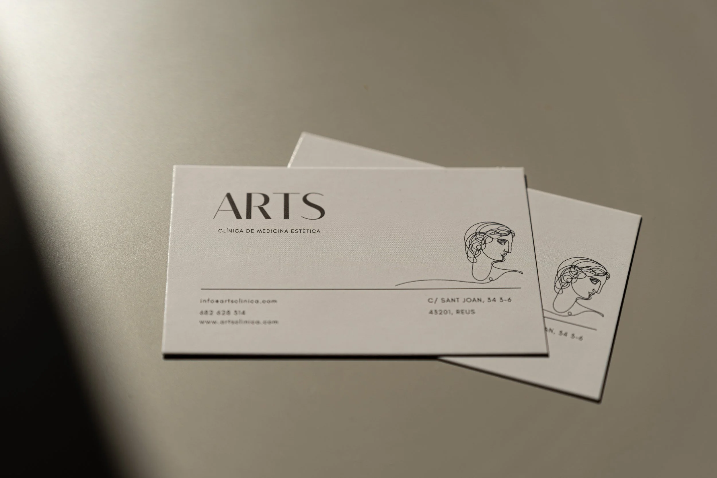 Tarjetas de visita en color blanco con ilustraciones de perfiles de rostro femenino y texto que dice 'ARTS, Clínica de Medicina Estética, dirección, teléfono y sitio web' sobre una superficie gris clara.