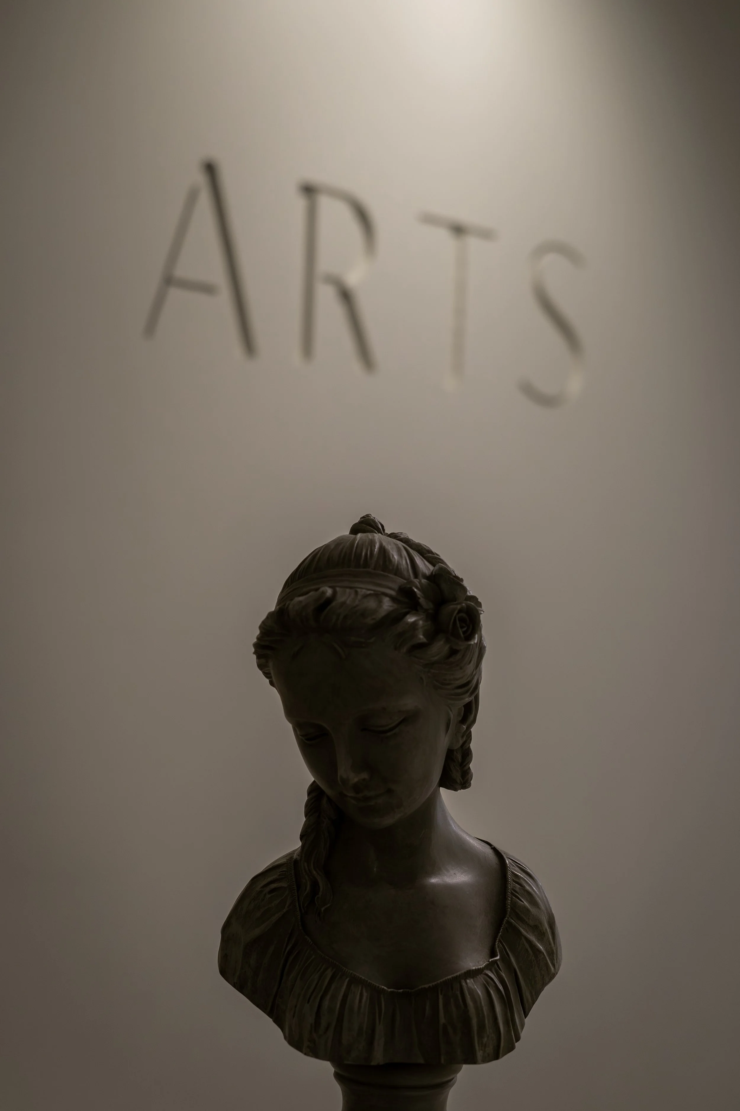 Escultura de busto de mujer con cabello trenzado, vista desde abajo, con letras 'ARTS' en la pared en el fondo