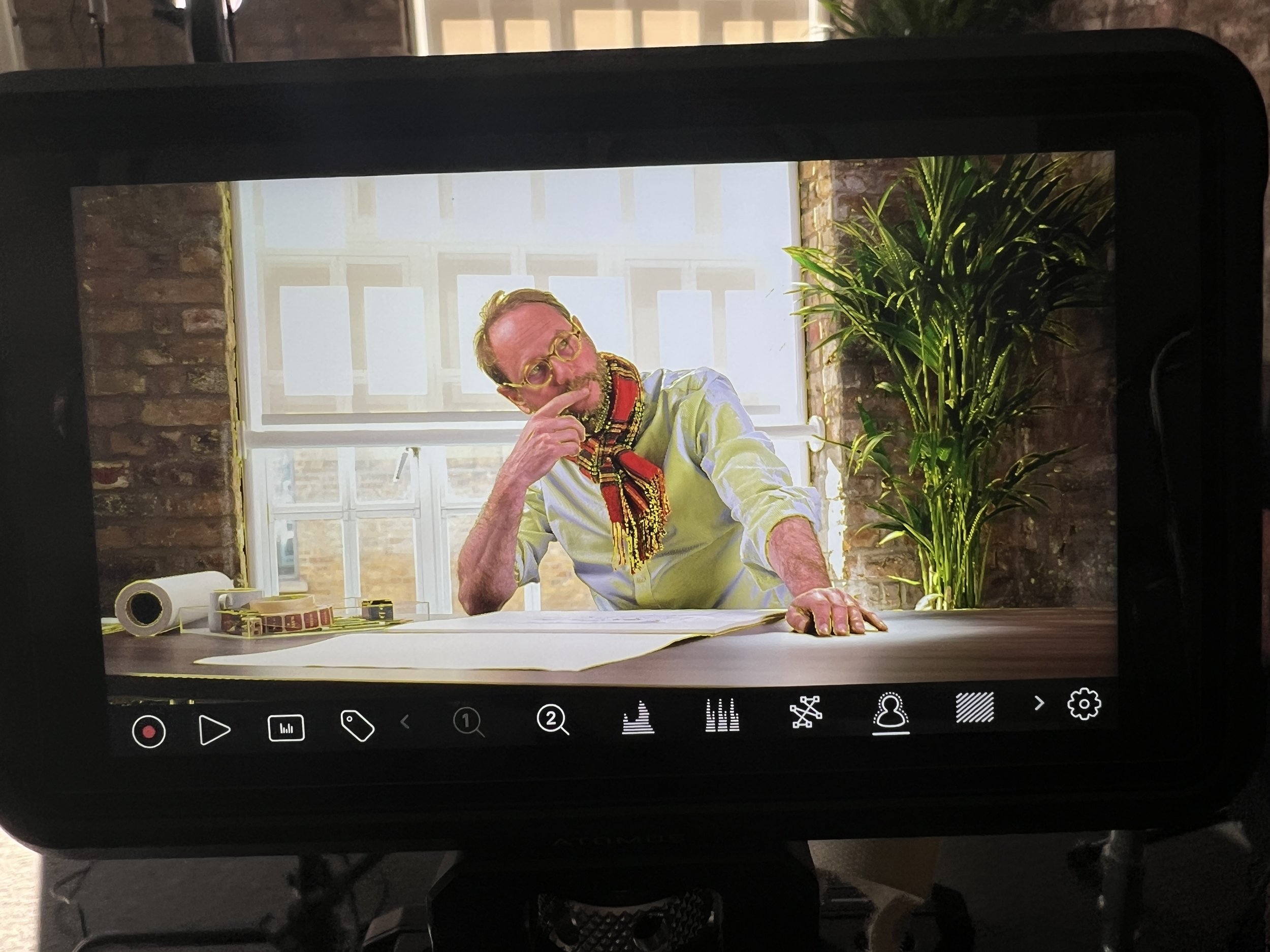 A man with glasses and a colorful scarf sitting at a desk in front of a large window, with a potted plant on the side and office supplies on the desk.
