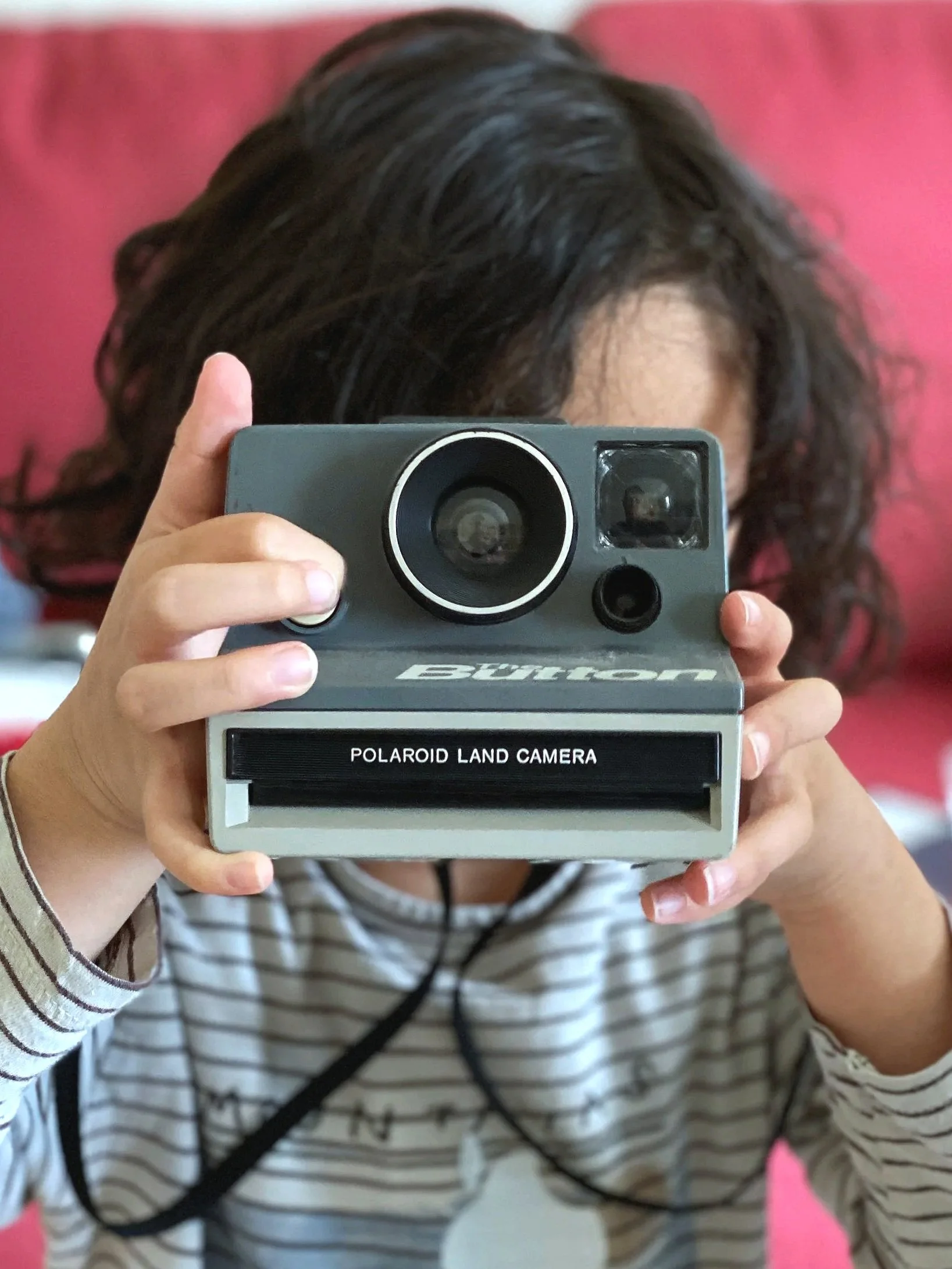 Child holding a gray Polaroid Land Camera with the lens pointed forward, partially obscuring their face, against a red background.