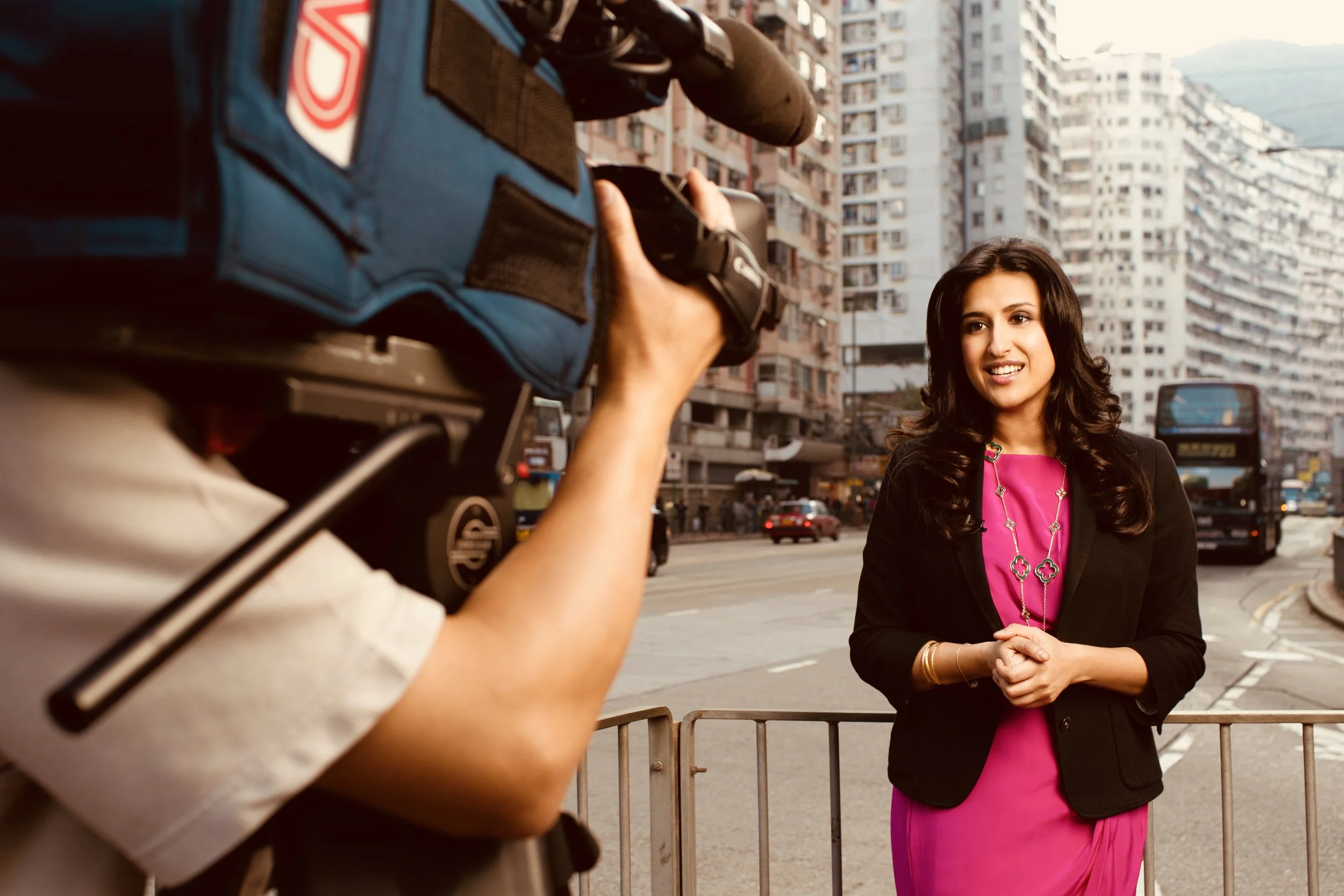A woman in a pink dress and black blazer being interviewed on a city street with tall buildings and a bus in the background.