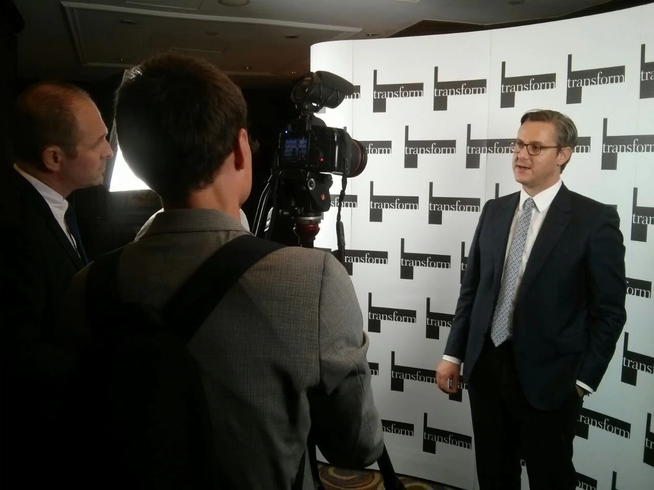 A man in a suit and glasses being interviewed or giving a statement in front of a backdrop with the word 'transform' repeated on it. Two other men are filming or recording him with a camera.