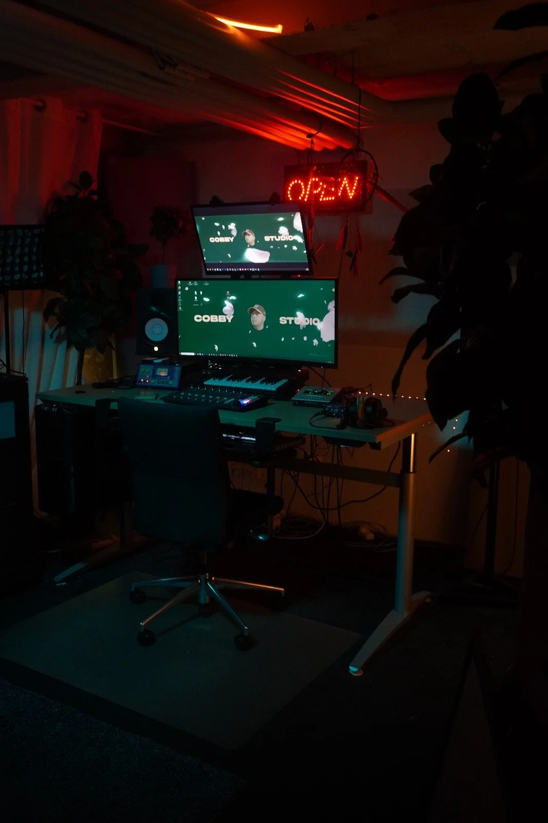 Ein Heimstudio mit zwei Computermonitoren, einem Keyboard, Lautsprechern und verschiedenen Musikinstrumenten, Beleuchtung durch eine rote Neon-Open-Leuchttafel, Pflanzen und diverse Geräte auf dem Tisch, dunkle Atmosphäre.