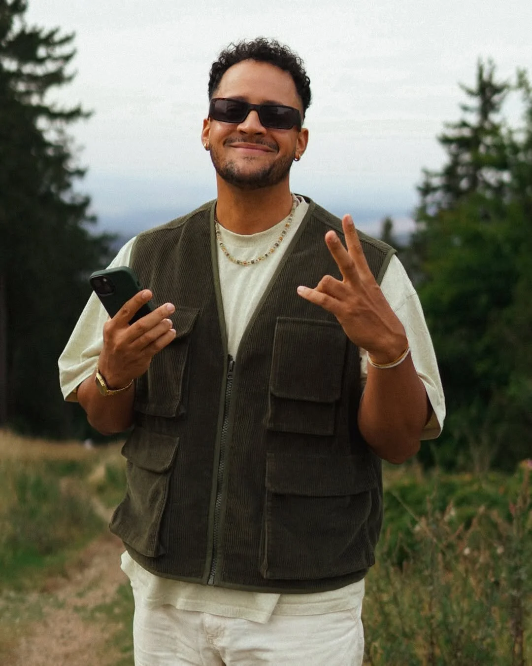 Lächelnder Mann mit Sonnenbrille, Kette und Armbändern, der eine Hand mit Peace-Zeichen zeigt und in der anderen ein Smartphone hält, im Freien mit Bäumen im Hintergrund.