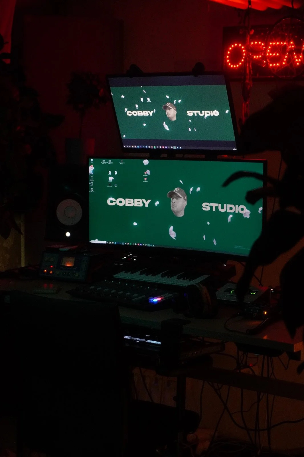 Zwei Computer-Monitore, ein Keyboard, ein Lautsprecher, ein Mischpult und anderes Audio-Equipment in einem dunklen Raum. Auf den Monitoren ist ein grüner Hintergrund mit einem Gesicht in der Mitte, umgeben von weißen Schriftzügen ‚COBBY STUPID‘ und weißen Federn.