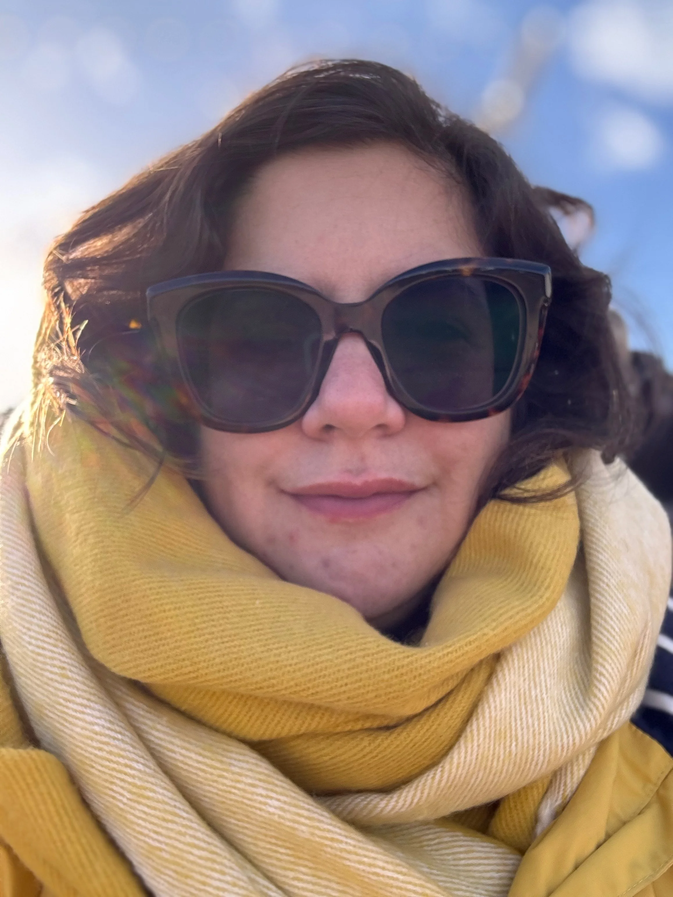 Close-up of a woman with curly brown hair wearing large dark sunglasses and a yellow scarf outdoors.