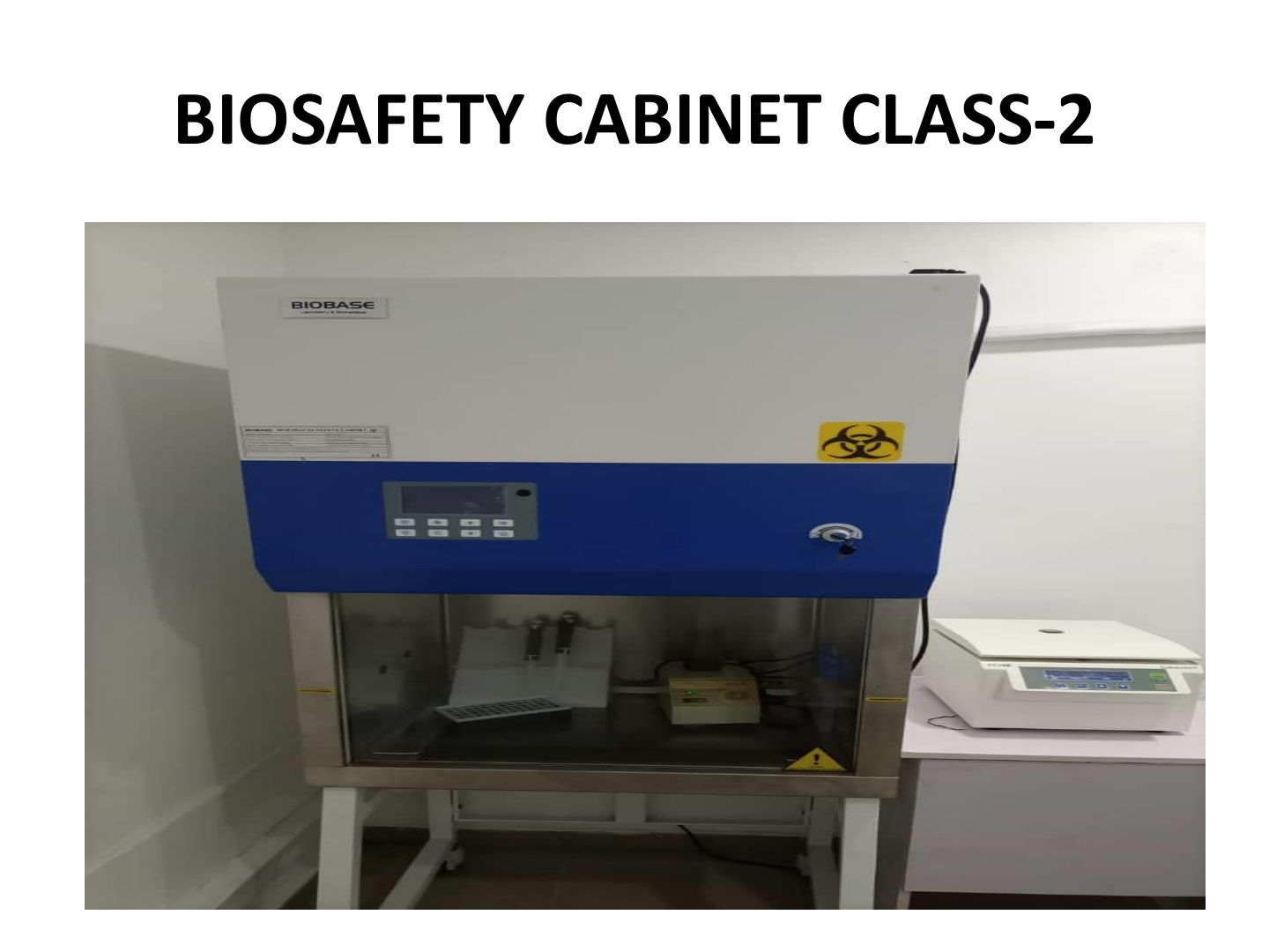 Biosafety cabinet Class 2 in a laboratory setting, with a digital control panel and biohazard warning label, accompanied by a smaller machine on the right.