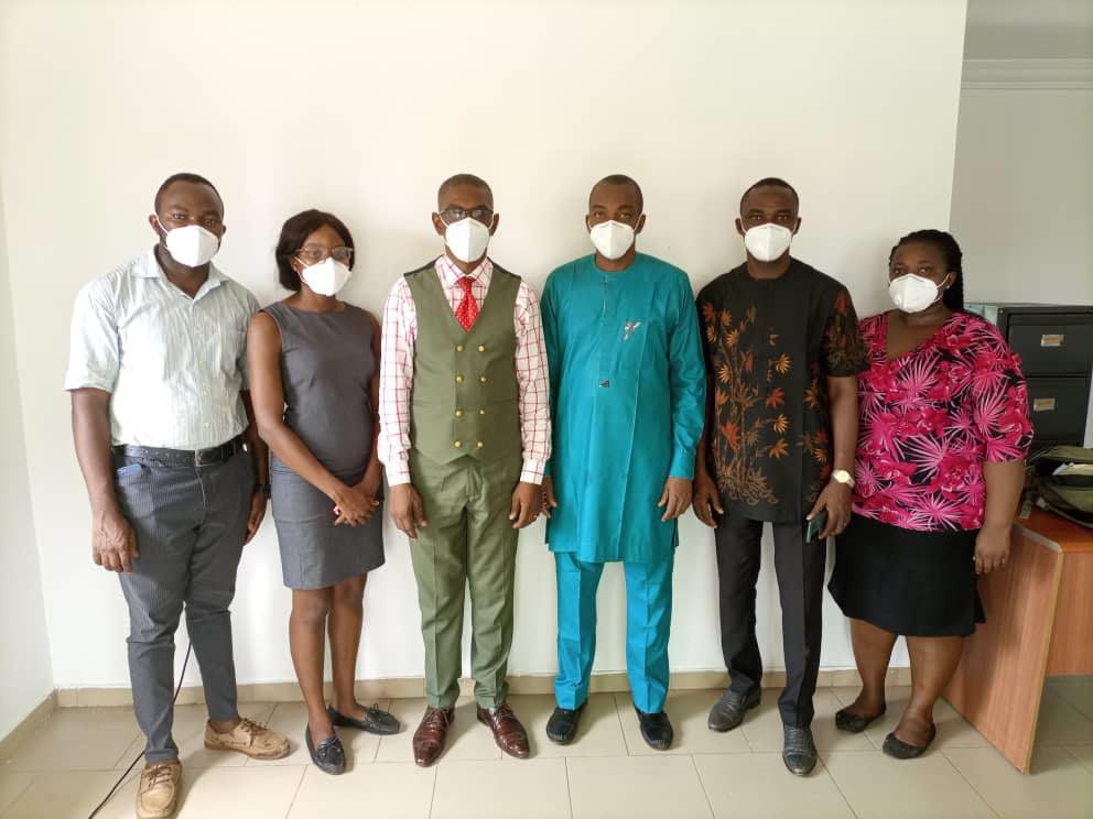Six people standing in a row, all wearing face masks, indoors against a plain wall.