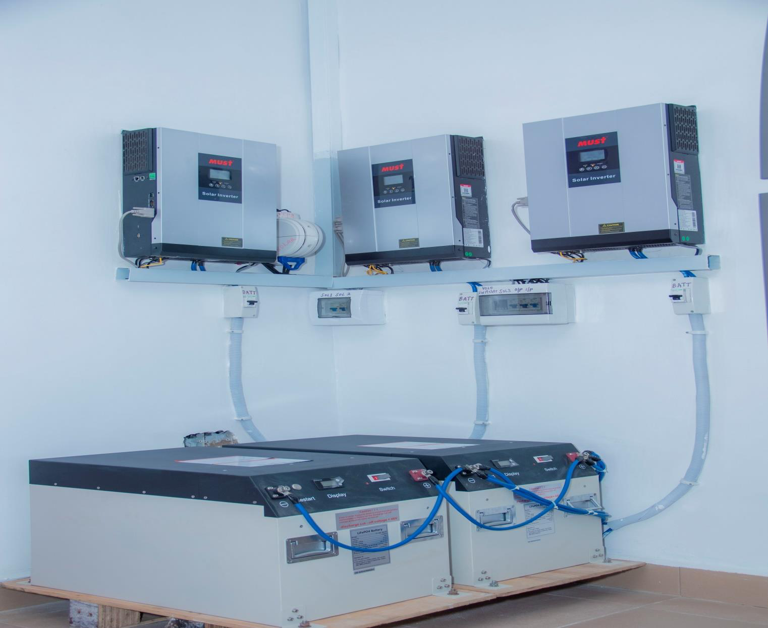 Three gray solar inverters mounted on a white wall with electrical wiring, connected to a larger white battery storage system below.