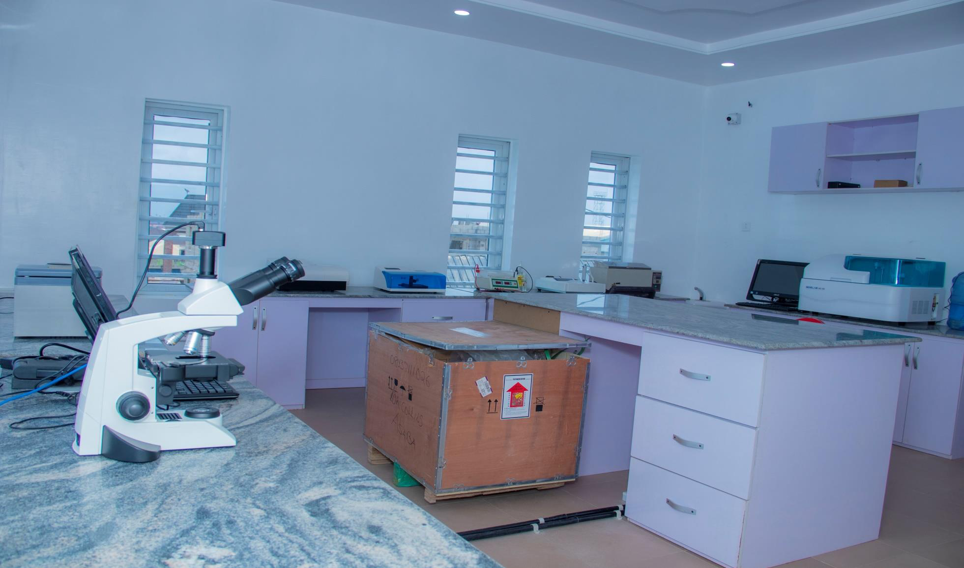 Laboratory room with scientific equipment including a microscope, computer, and various devices on the countertop, a large cardboard box in the center, and windows with metal bars letting in natural light.