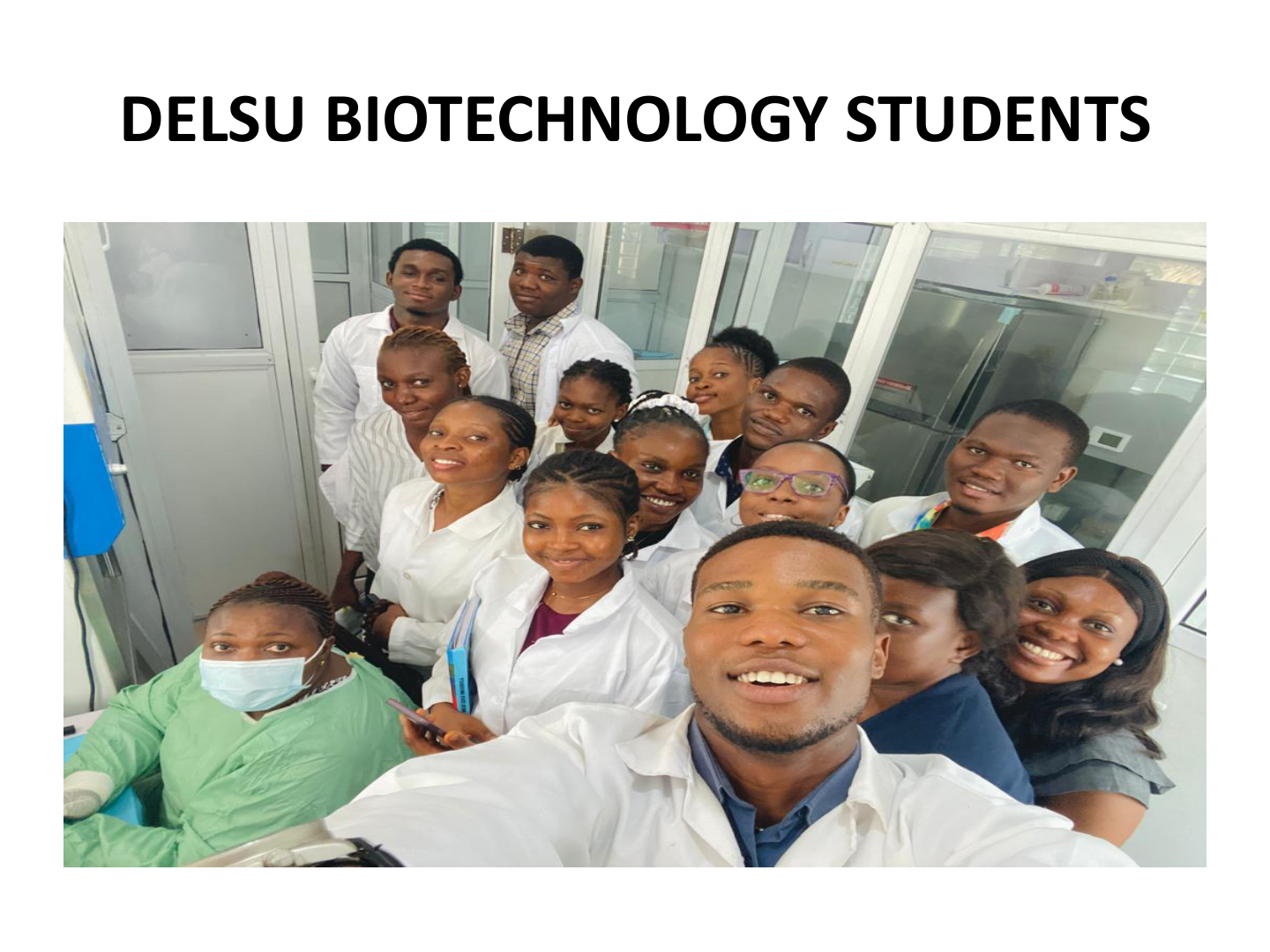 Group of students and instructors in a laboratory, some wearing white lab coats and face masks, taking a selfie together