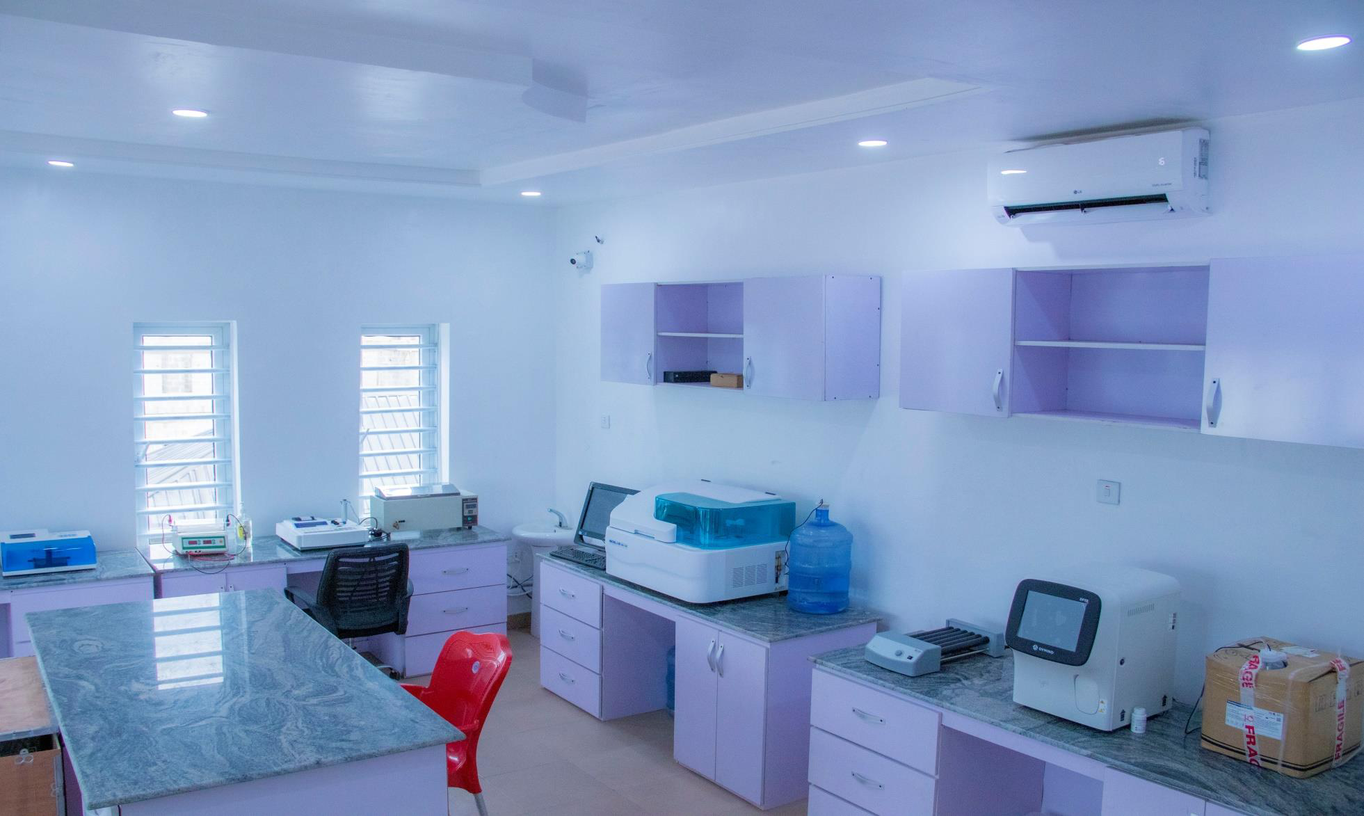 A medical laboratory with white walls, purple cabinets, and various laboratory equipment on countertops, including centrifuges and other devices; a red chair is in the foreground.