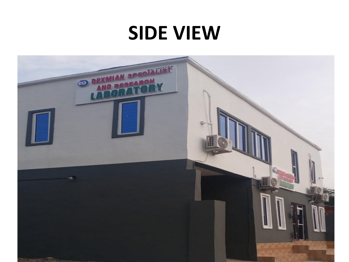 Side view of a white two-story building with blue and green window frames and multiple air conditioning units. A sign on the building reads 'Dexmian Specialist and Research Laboratory'.
